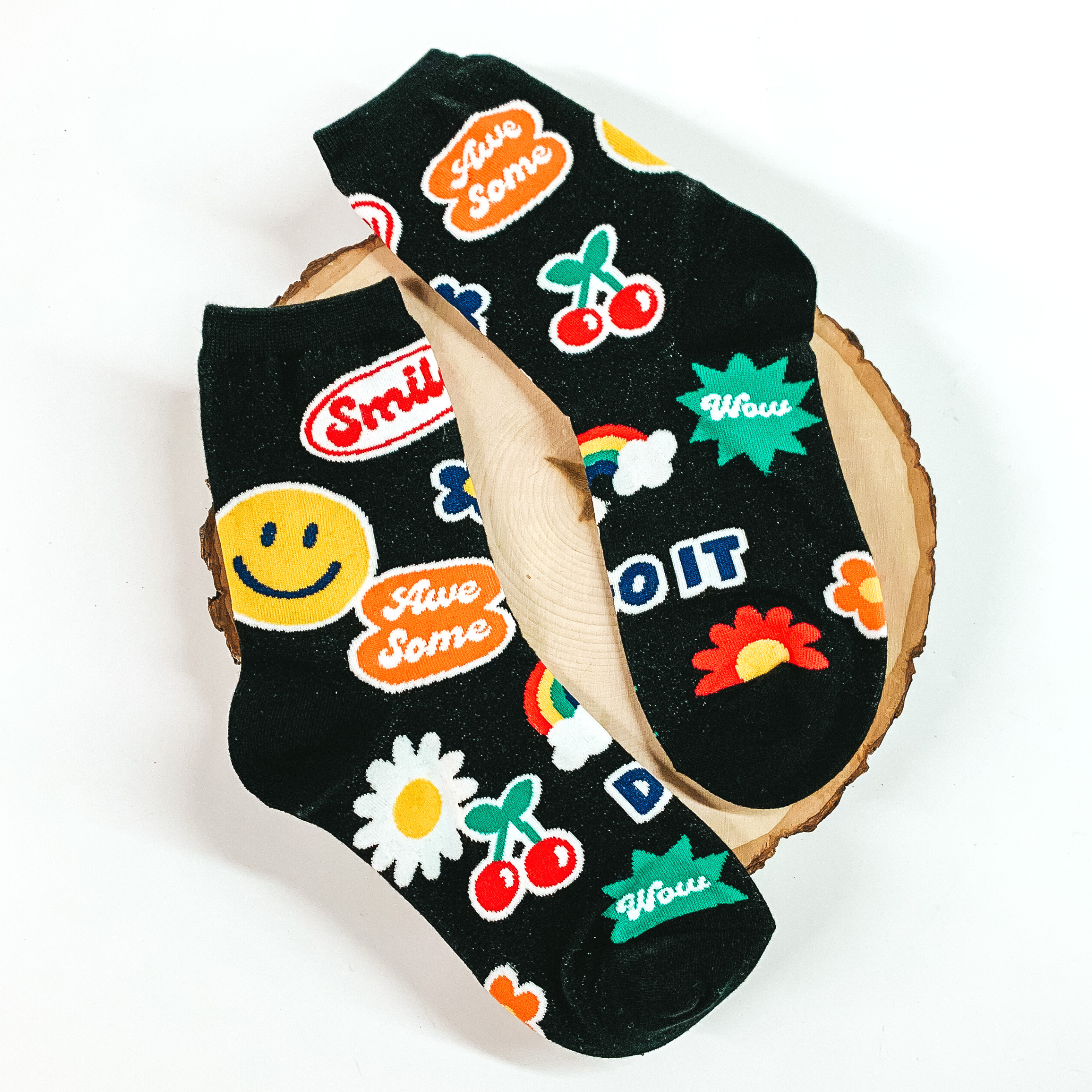 Black ankle socks with a retro, sticker design. These stickers include a smiley face, cherries, flowers, rainbow, and words. These socks are pictured on a piece of wood on a white background. 
