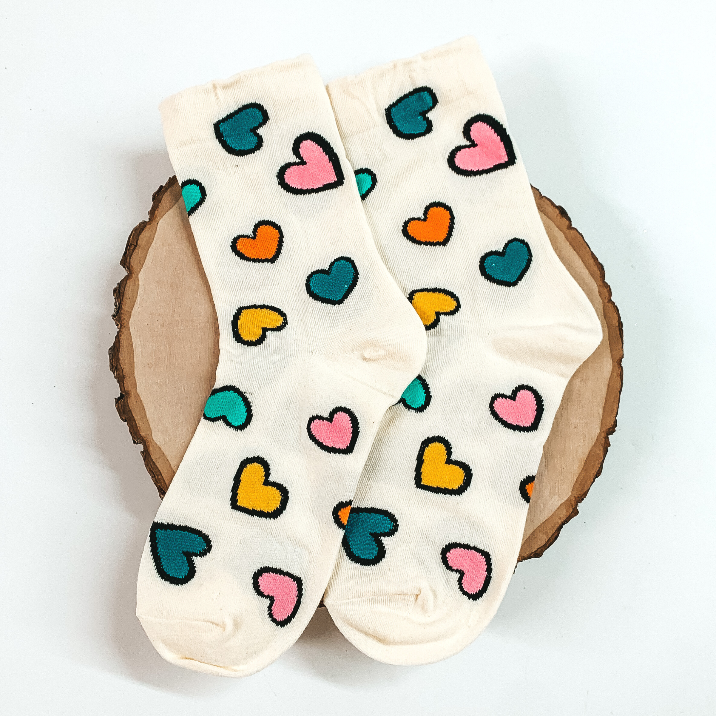 Cream ankle socks with a multicolored heart design. The hearts have a black outline. These socks are pictured on a piece of wood on a white background. 