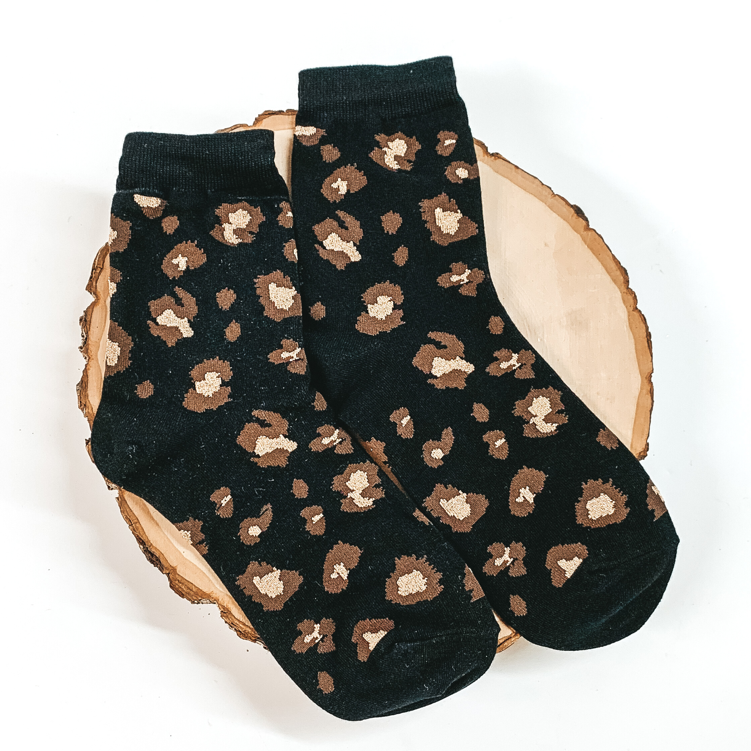 Black ankle socks with a brown and champagne leopard print. These socks are pictured on a piece of wood on a white background. 