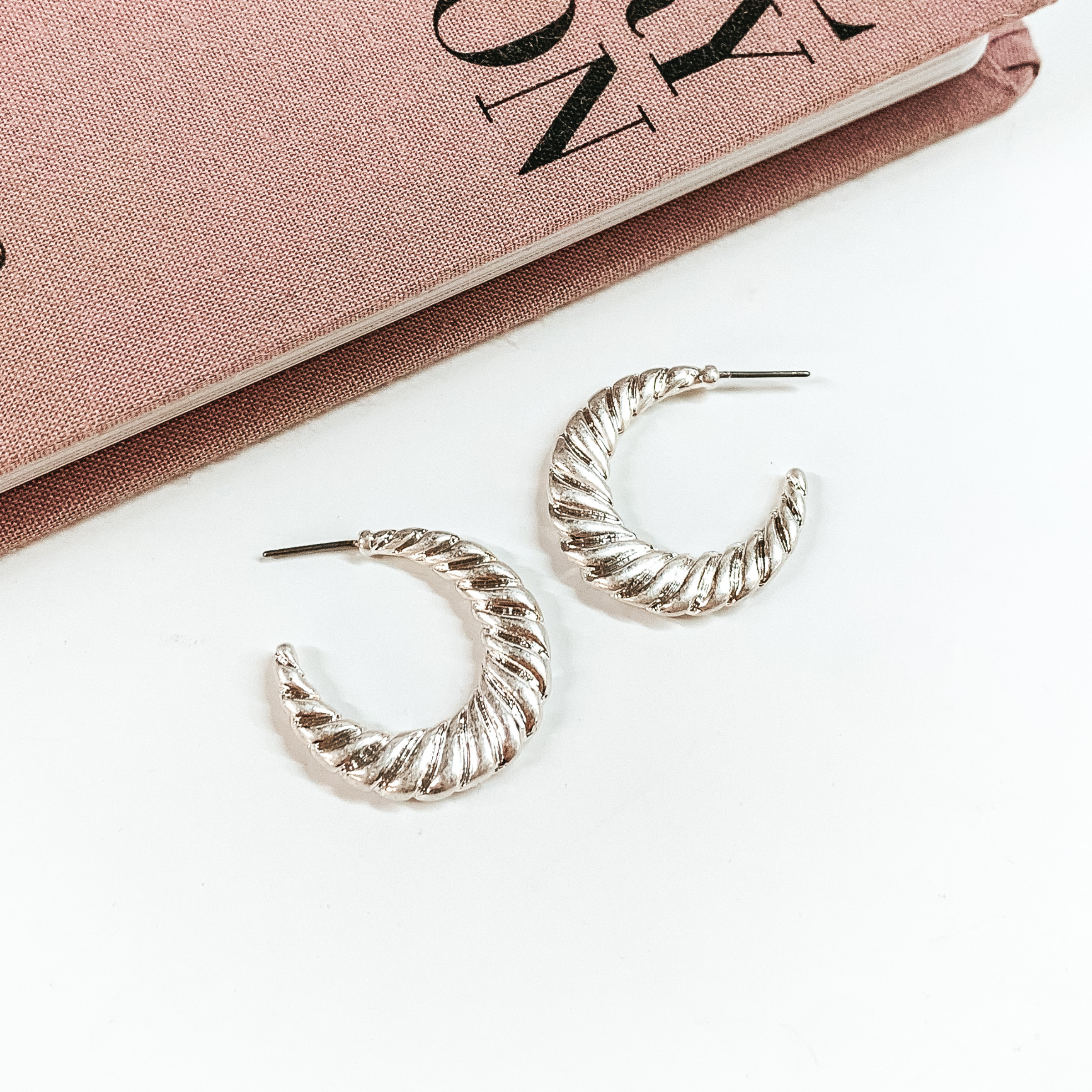 Silver, twisted hoop earrings pictured on a white background with a mauve colored book at the top of the picture. 