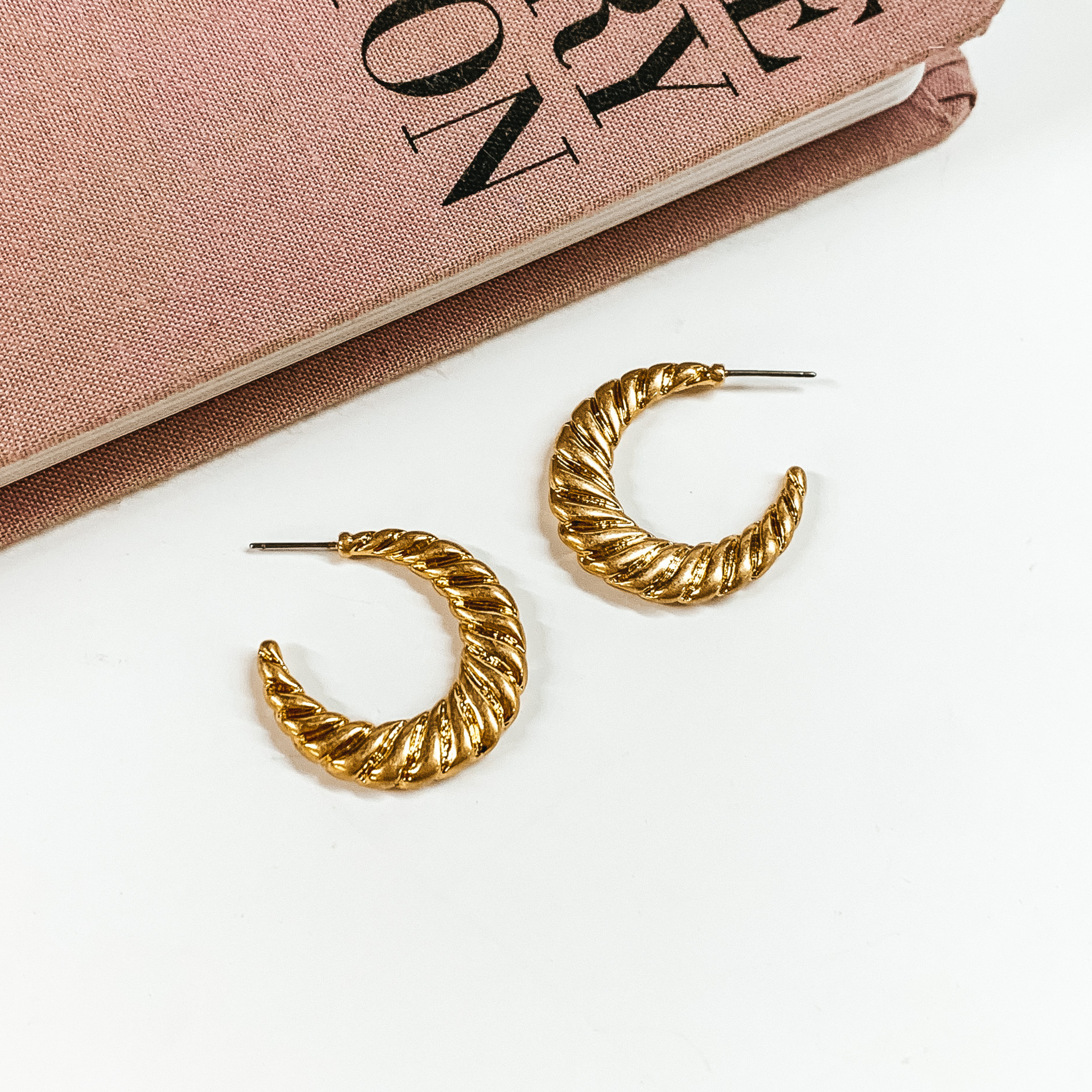 Gold, twisted hoop earrings pictured on a white background with a mauve colored book at the top of the picture. 