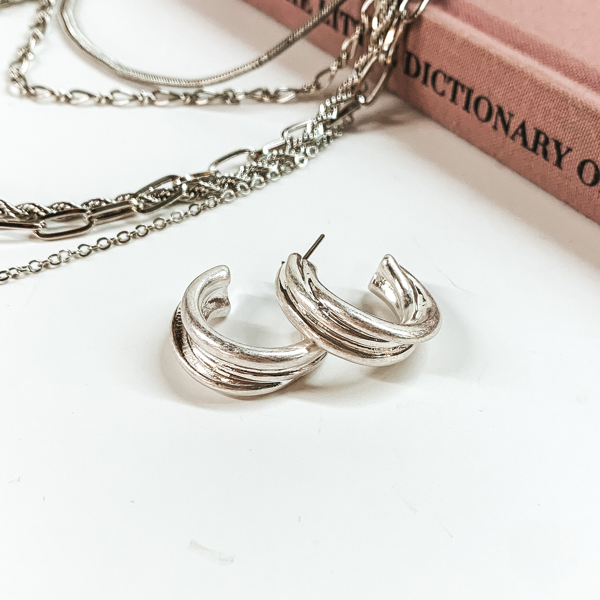 Silver, layered hoop earrings pictured on a white background with a mauve colored book and silver chains at the top of the picture. 
