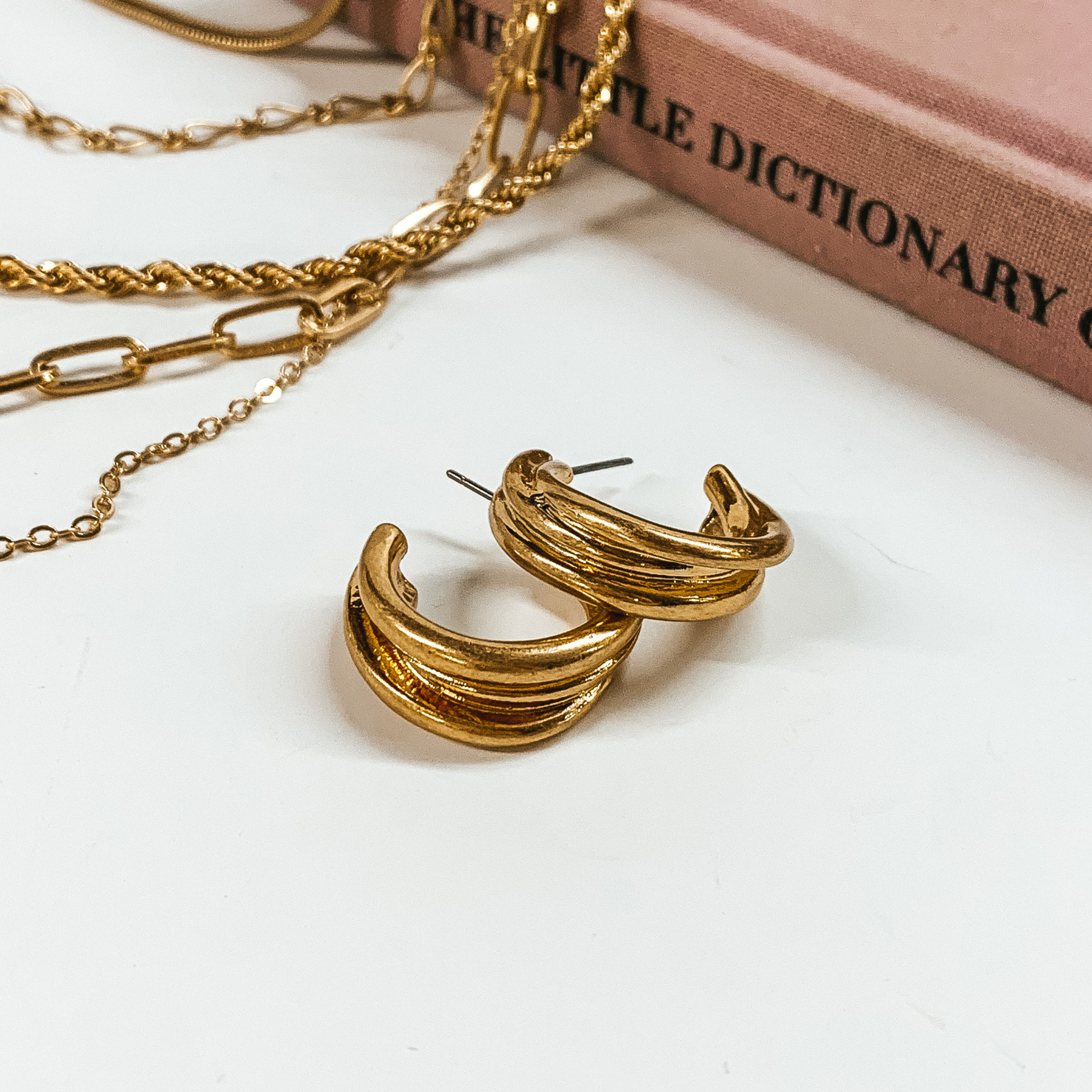 Gold, layered hoop earrings pictured on a white background with a mauve colored book and silver chains at the top of the picture. 