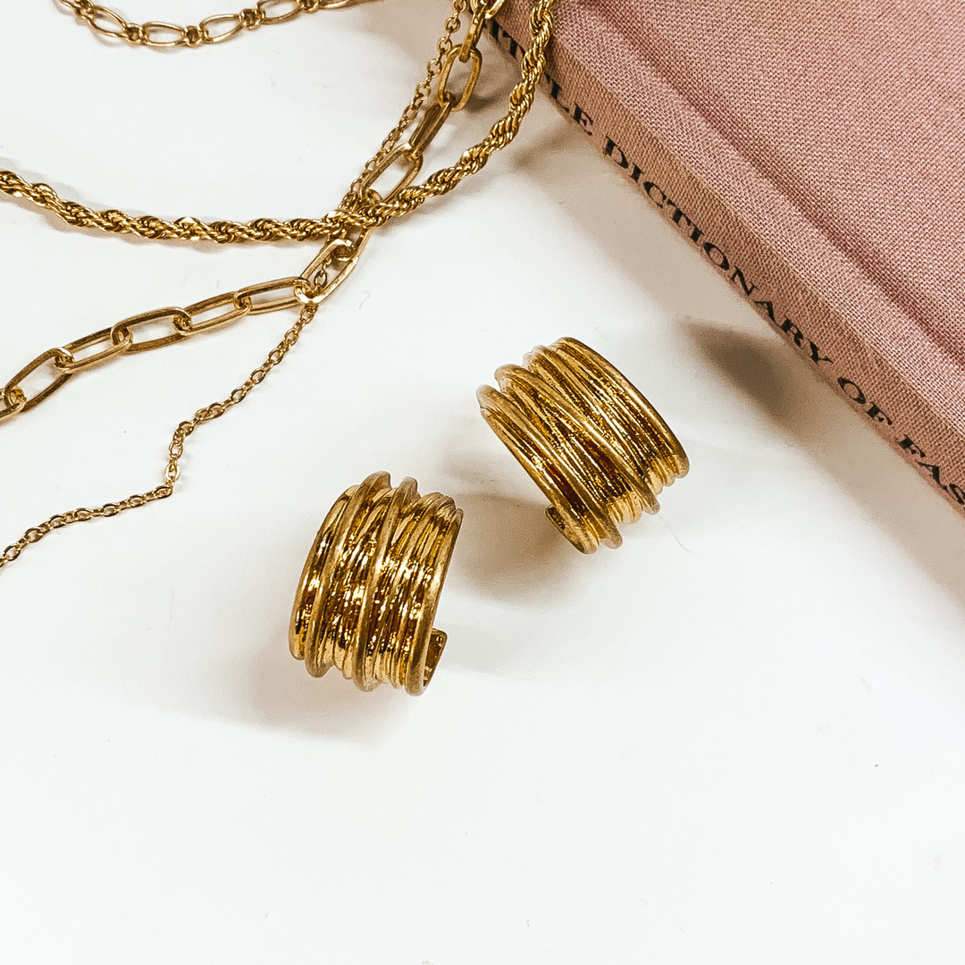 Gold, thick hoop earrings pictured on a white background with a mauve colored book and silver chains at the top of the picture. 