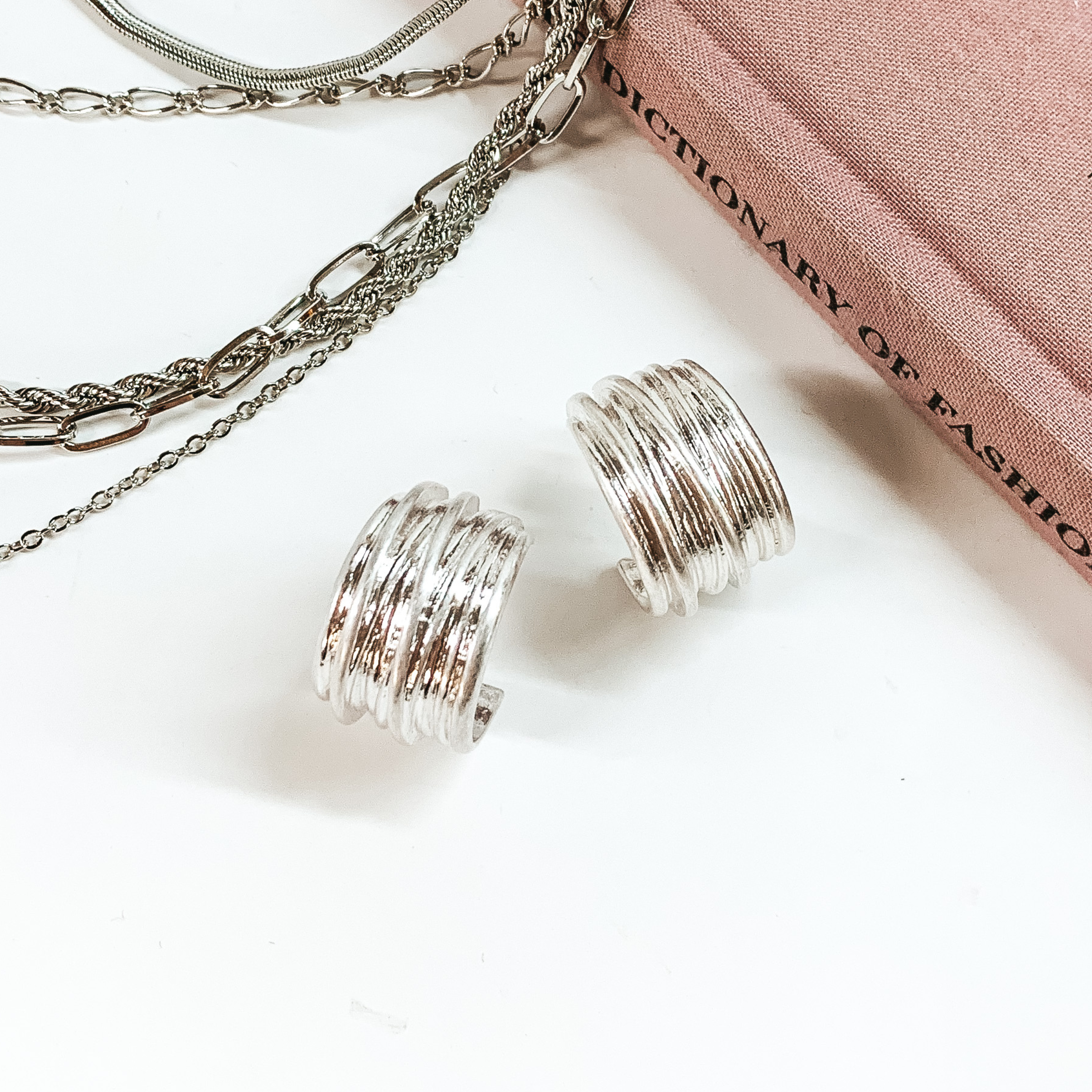 Silver, thick hoop earrings pictured on a white background with a mauve colored book and silver chains at the top of the picture. 