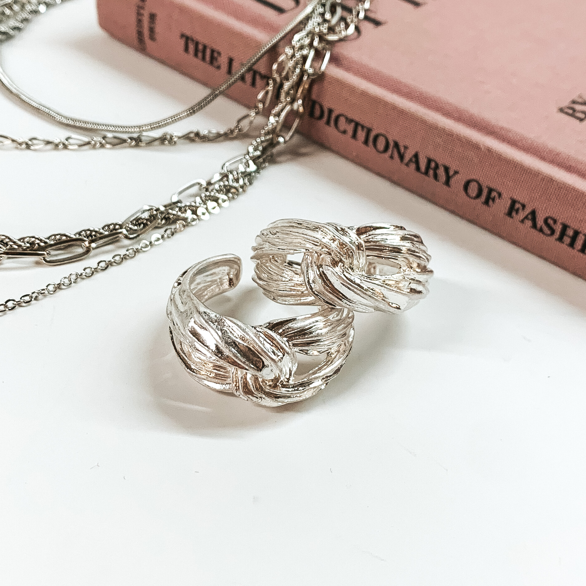 Silver, knot hoop earrings pictured on a white background with a mauve book and silver chains in the back of the picture. 