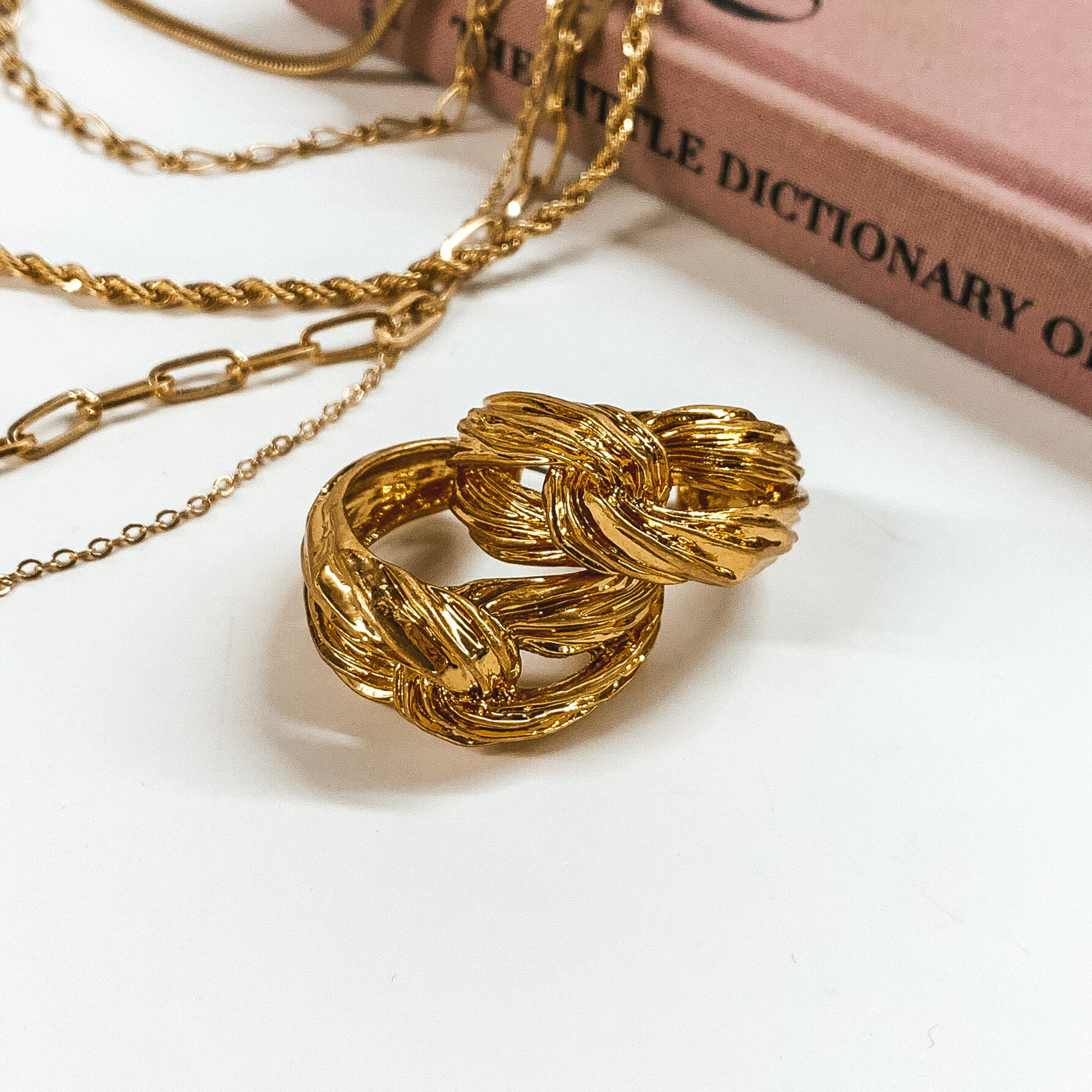 Gold, knot hoop earrings pictured on a white background with a mauve book and gold chains in the back of the picture. 
