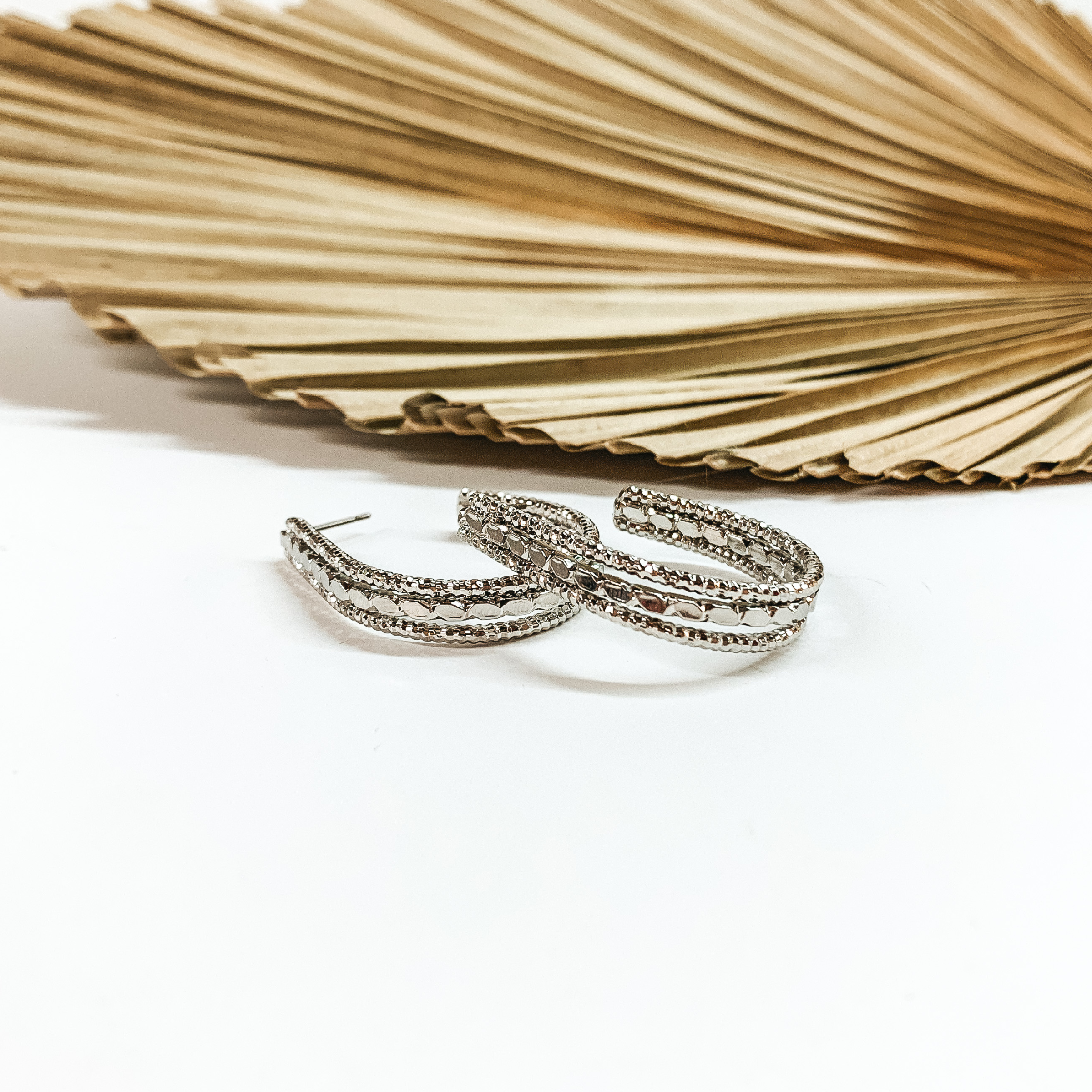 Teardrop, silver hoop earrings with different textures. These earrings are pictured in front a sage green leaf on a white background. 