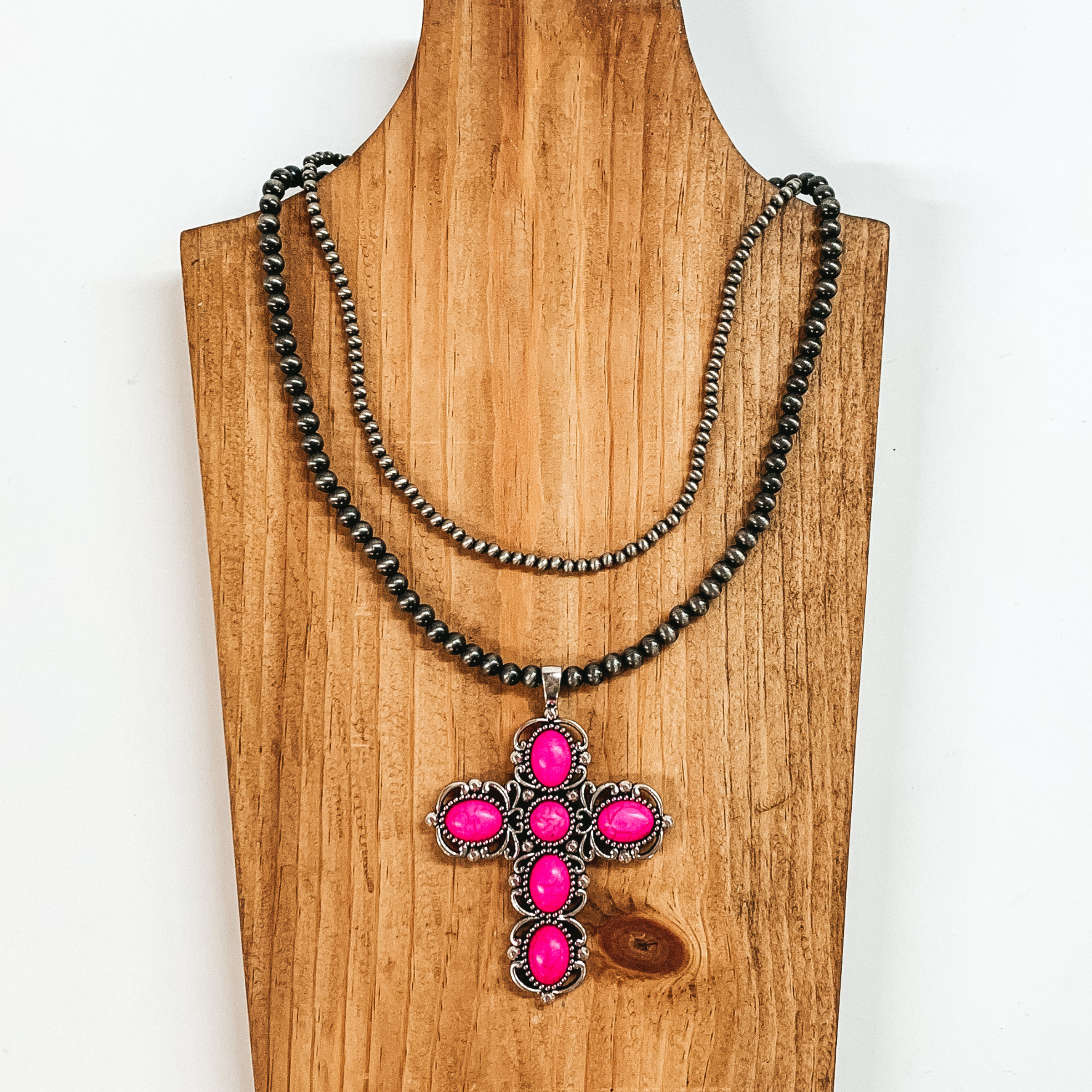 Two strand neckace with different sized silver beaded strands of different lengths with a cross pendant that has pink stones. This necklace is pictured on a wooden necklace holder on a white background. 