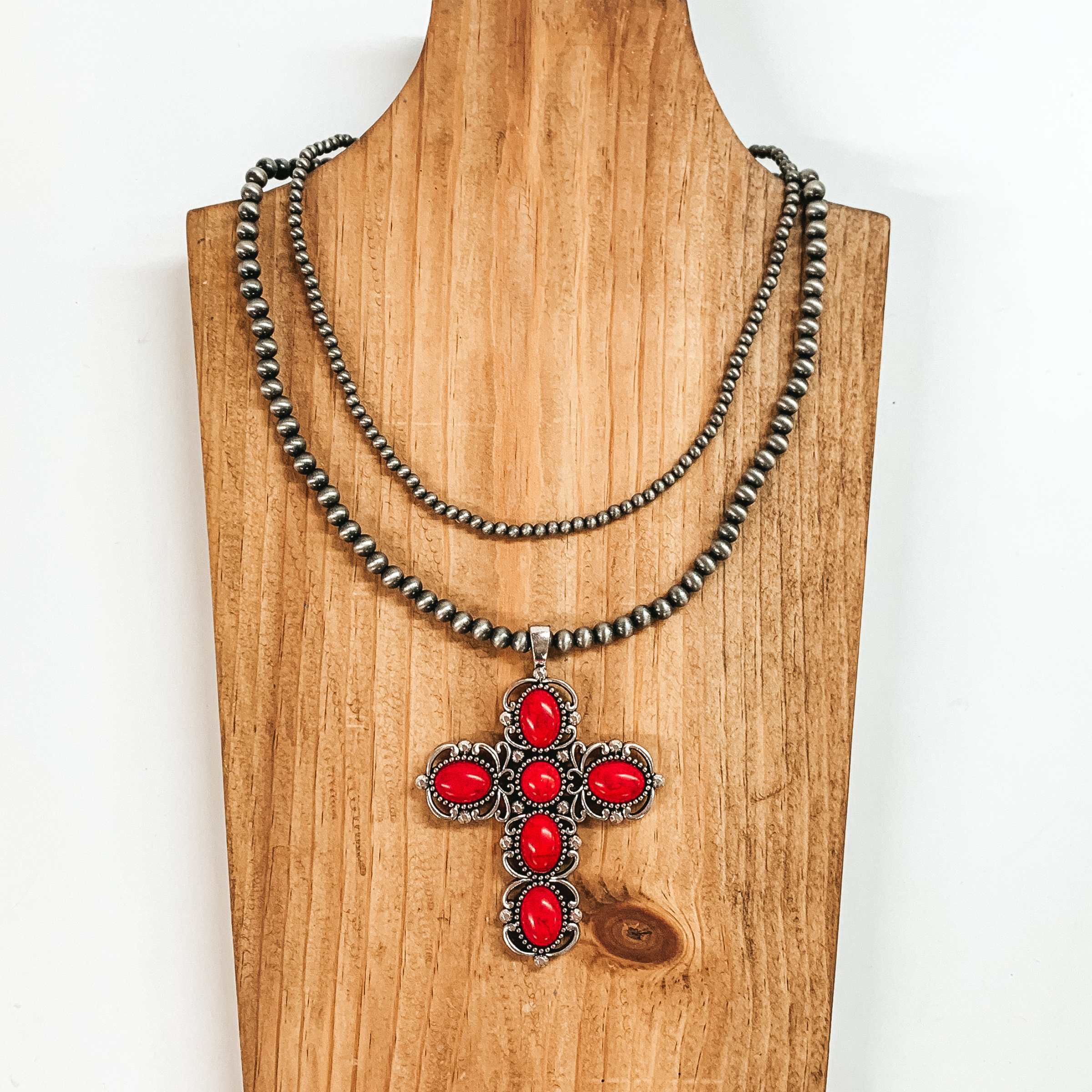 Two strand neckace with different sized silver beaded strands of different lengths with a cross pendant that has red stones. This necklace is pictured on a wooden necklace holder on a white background. 