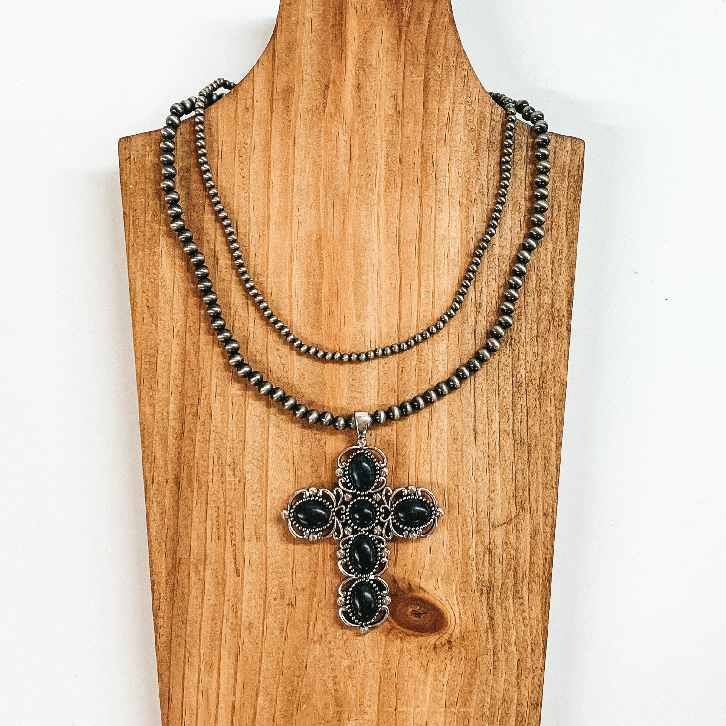 Two strand neckace with different sized silver beaded strands of different lengths with a cross pendant that has black stones. This necklace is pictured on a wooden necklace holder on a white background. 