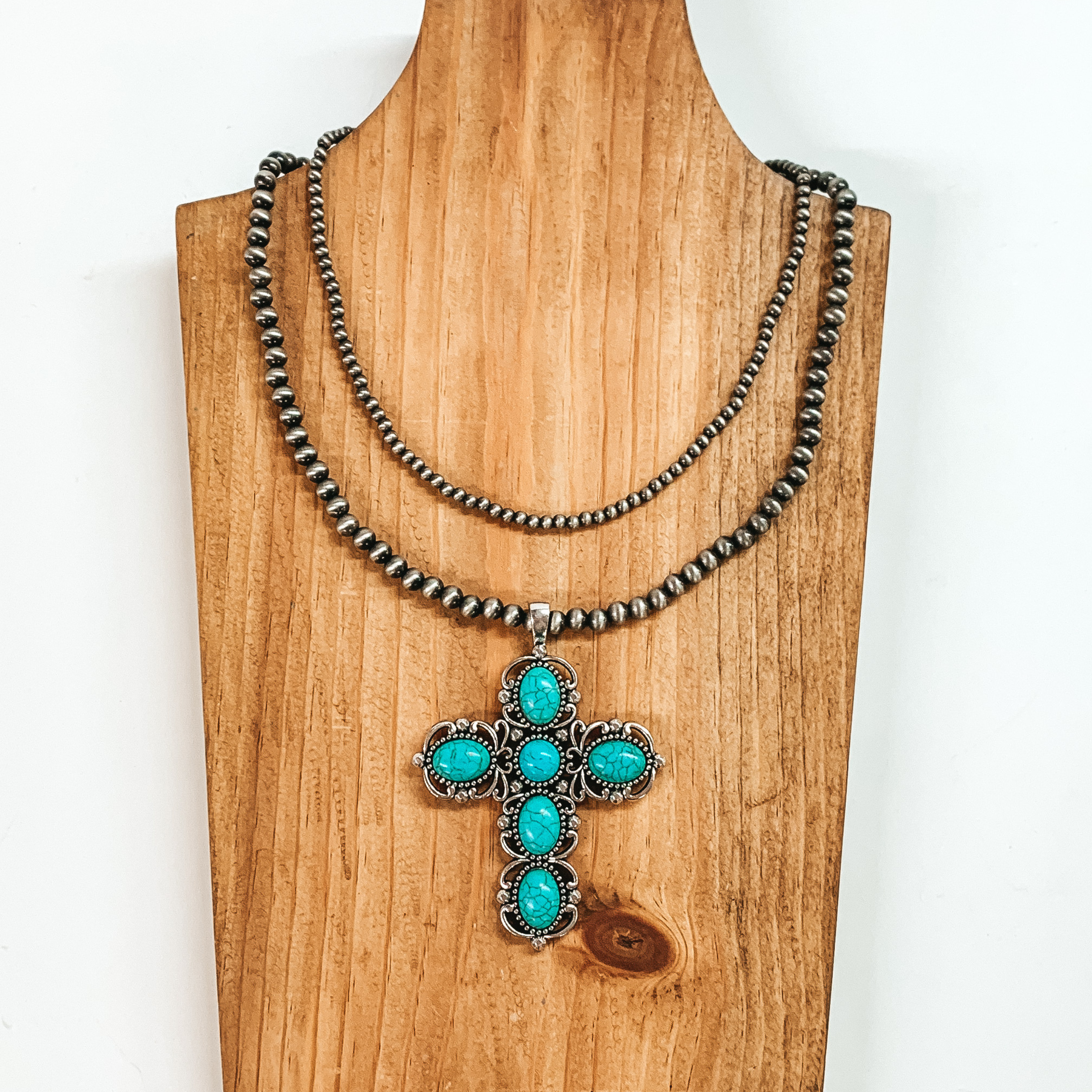 Two strand neckace with different sized silver beaded strands of different lengths with a cross pendant that has turquoise stones. This necklace is pictured on a wooden necklace holder on a white background. 