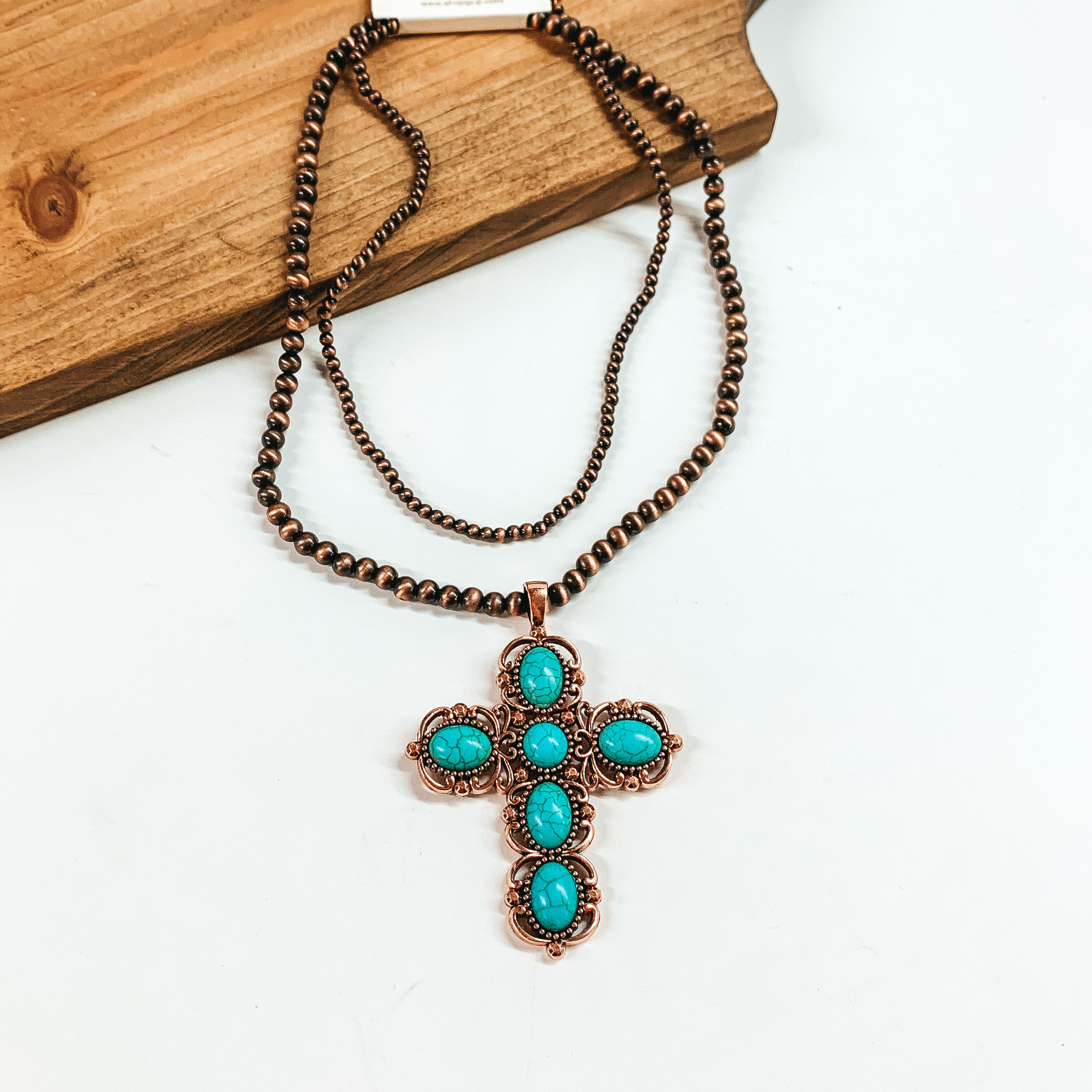 Two strand neckace with different sized copper beaded strands of different lengths with a cross pendant that has turquoise stones. This necklace is pictured on a white background. 