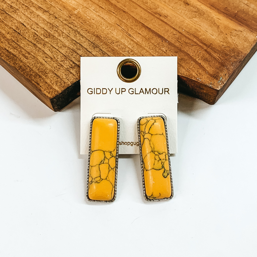 Rectangle, yellow stone earrings with a thin, silver outline. These earrings are pictured in front of a brown block on a white background. 