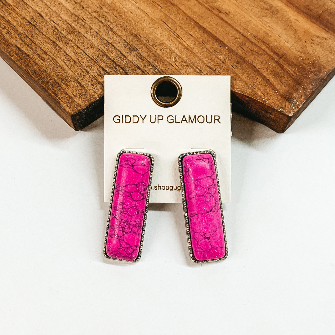 Rectangle, pink stone earrings with a thin, silver outline. These earrings are pictured in front of a brown block on a white background. 