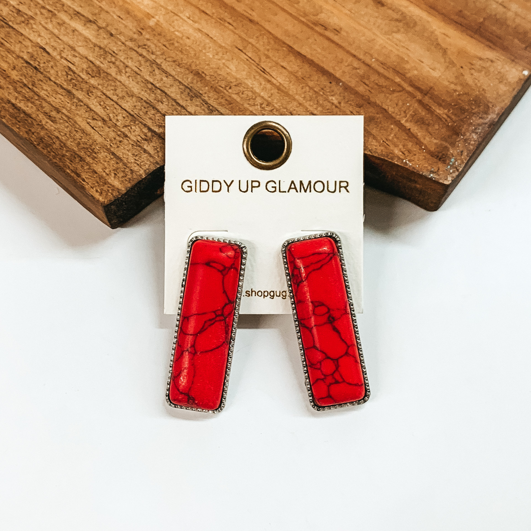 Rectangle, red stone earrings with a thin, silver outline. These earrings are pictured in front of a brown block on a white background. 
