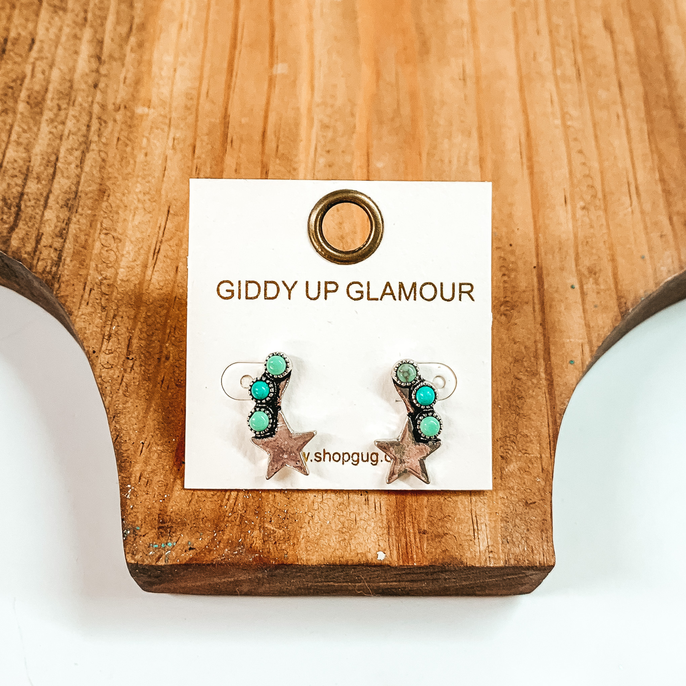 These earrings include three turquoise stones with a silver star at the bottom. These earrings are pictured on top of a brown block on a white background. 