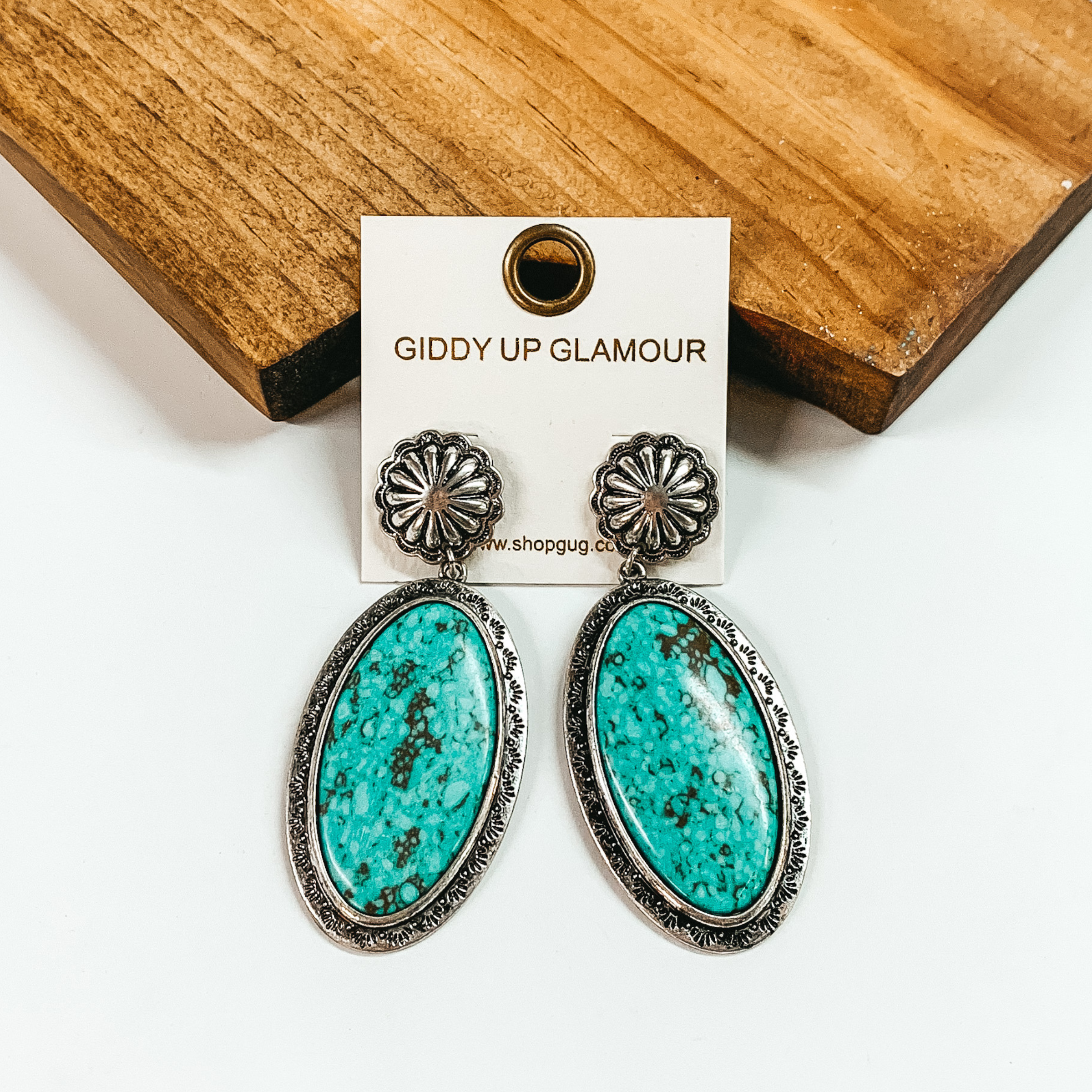Silver, circle concho earrings with an oval drop. The oval drop is a turquoise stone with a silver outline. These earrings are pictured in front of a brown block on a white background. 