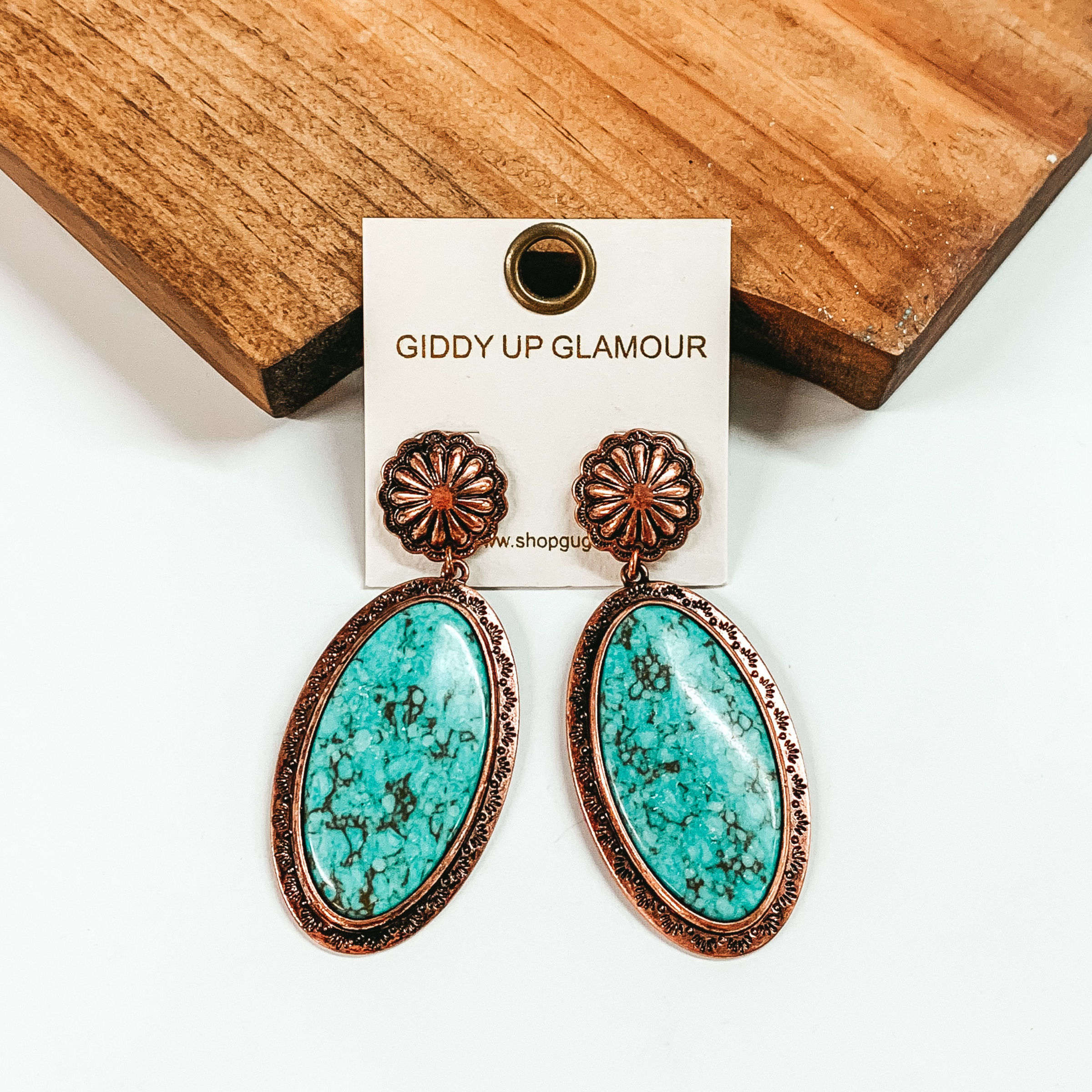Copper, circle concho earrings with an oval drop. The oval drop is a turquoise stone with a copper outline. These earrings are pictured in front of a brown block on a white background. 