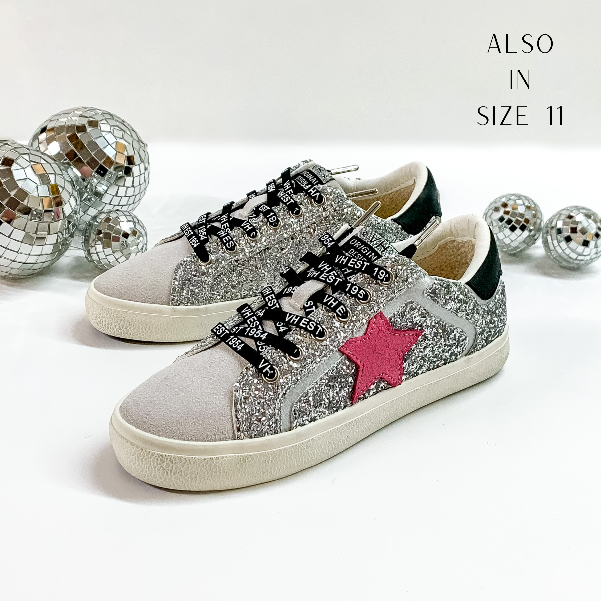 Grey colored tennis shoes and black shoelaces. The tennis shoes also have a pink fabric star patch on the side and silver glitter throughout. These shoes are pictured on a white background with disco balls behind them. 