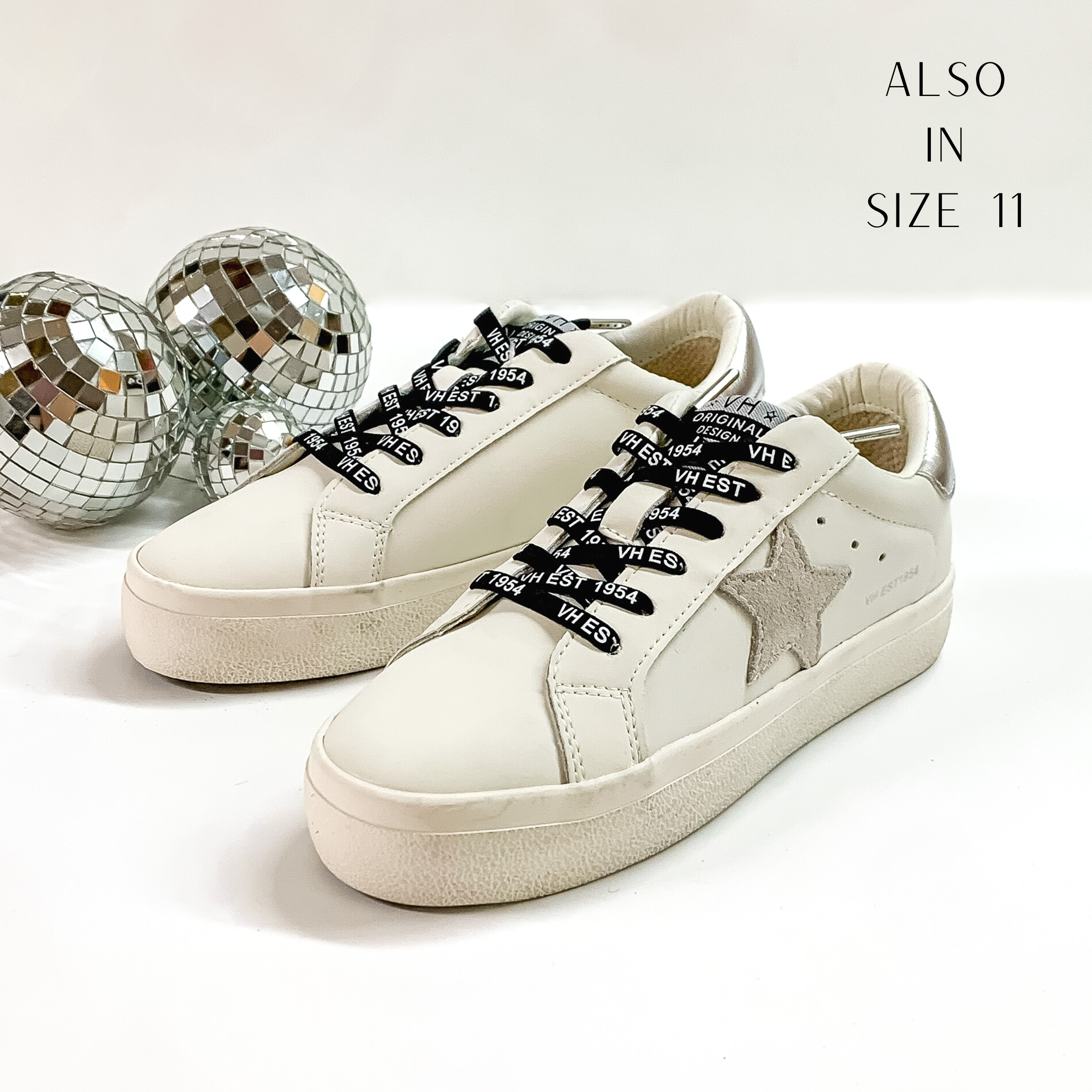 White tennis shoes and black shoelaces. The tennis shoes also have a silver glitter star patch on the side and a silver heel patch. These shoes are pictured on a white background with disco balls behind them. 