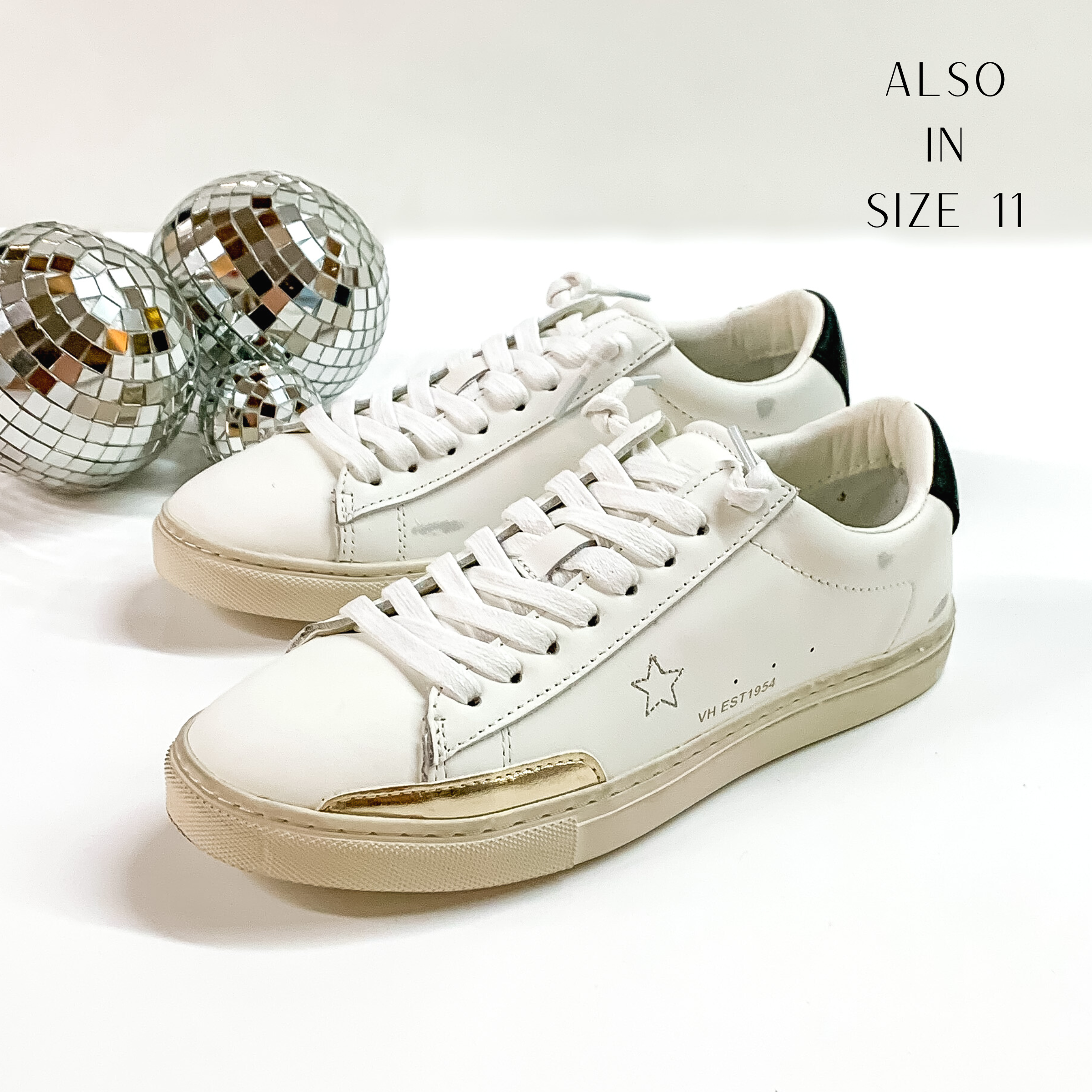 White tennis shoes and shoelaces. The tennis shoes also have a stitched star on the side, a black heel patch, and a gold outline on the side of the toe part. These shoes are pictured on a white background with disco balls behind them. 