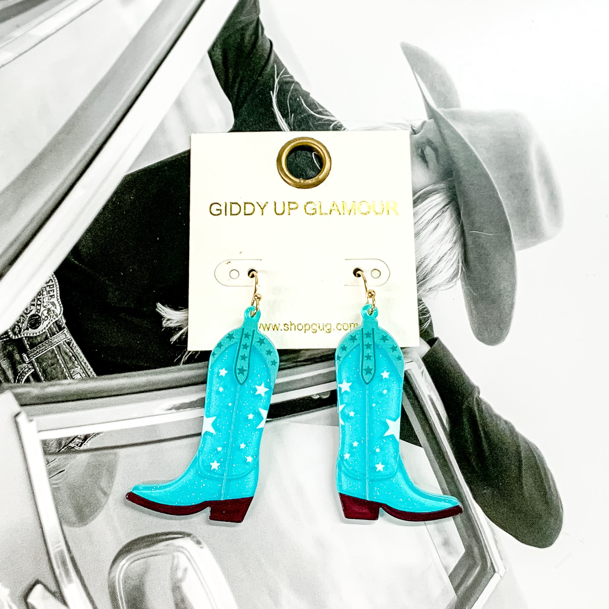 Cowboy boot dangle turquoise earrings pictured on a white earrings holder. These earrings have a star print and dark colored sole. These earrings are pictured on a black and white picture of a girl. 