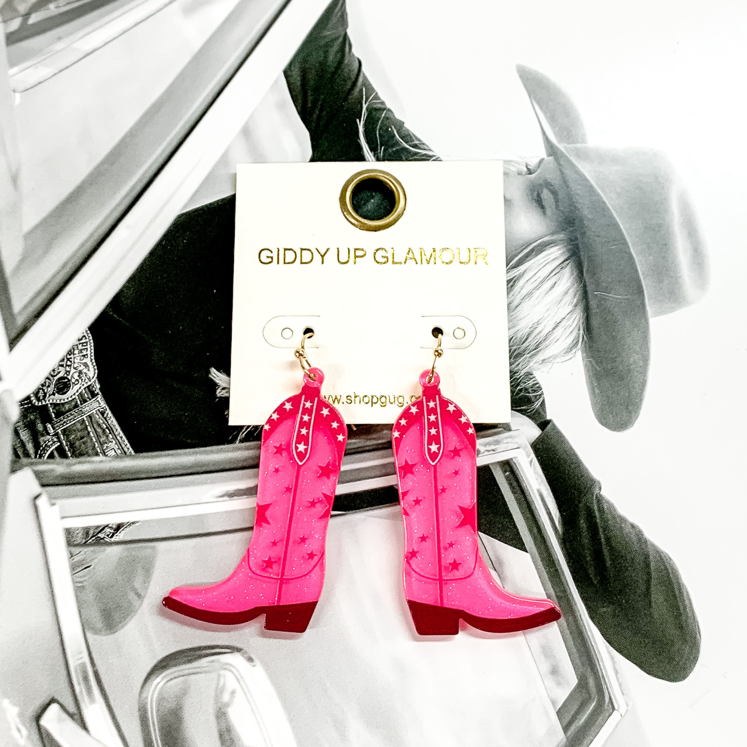 Cowboy boot dangle pink earrings pictured on a white earrings holder. These earrings have a star print and dark colored sole. These earrings are pictured on a black and white picture of a girl. 