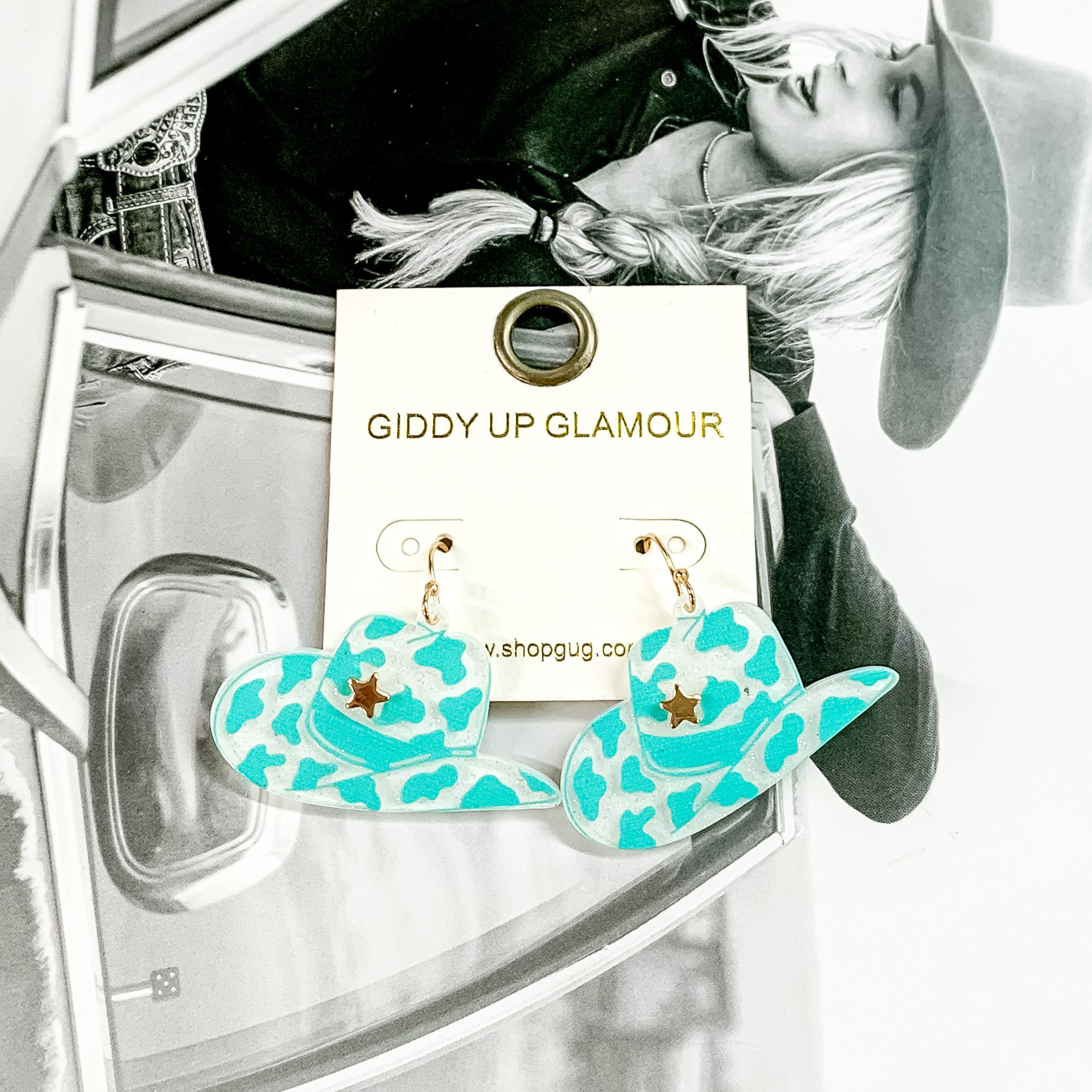 Cowboy hat dangle earrings pictured on a white earrings holder. These earrings have a turquoise cow print on a white, glitter background with a gold star on the front of the hat. These earrings are pictured on a black and white picture of a girl. 