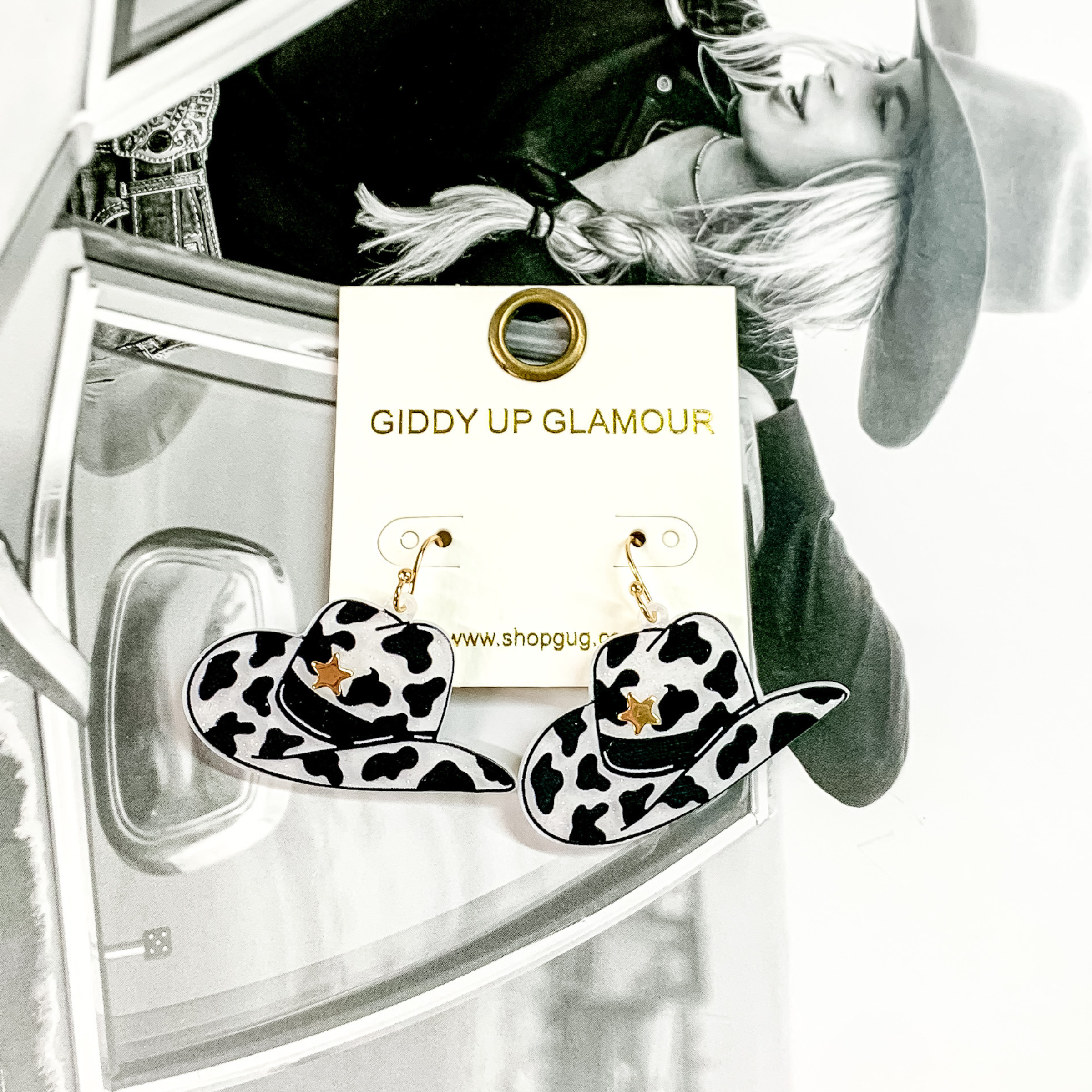 Cowboy hat dangle earrings pictured on a white earrings holder. These earrings have a black cow print on a white, glitter background with a gold star on the front of the hat. These earrings are pictured on a black and white picture of a girl. 
