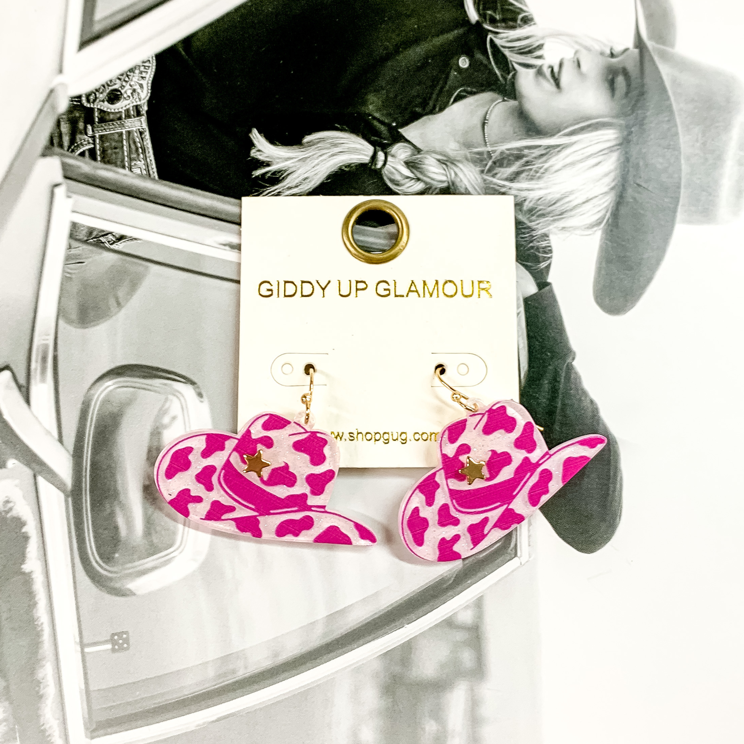 Cowboy hat dangle earrings pictured on a white earrings holder. These earrings have a fuchsia cow print on a white, glitter background with a gold star on the front of the hat. These earrings are pictured on a black and white picture of a girl. 