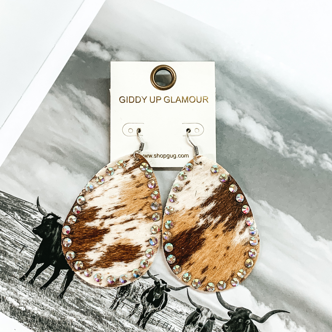 Teardrop dangle earrings that look like a white, tan, and brown cow print. These earrings also have an AB crystal outline. These earrings are pictured on a black and white picture of cows. 