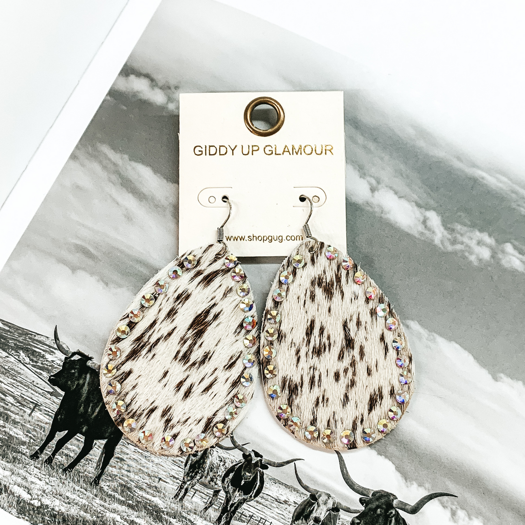 Teardrop dangle earrings that look like a white and brown cow print. These earrings also have an AB crystal outline. These earrings are pictured on a black and white picture of cows. 