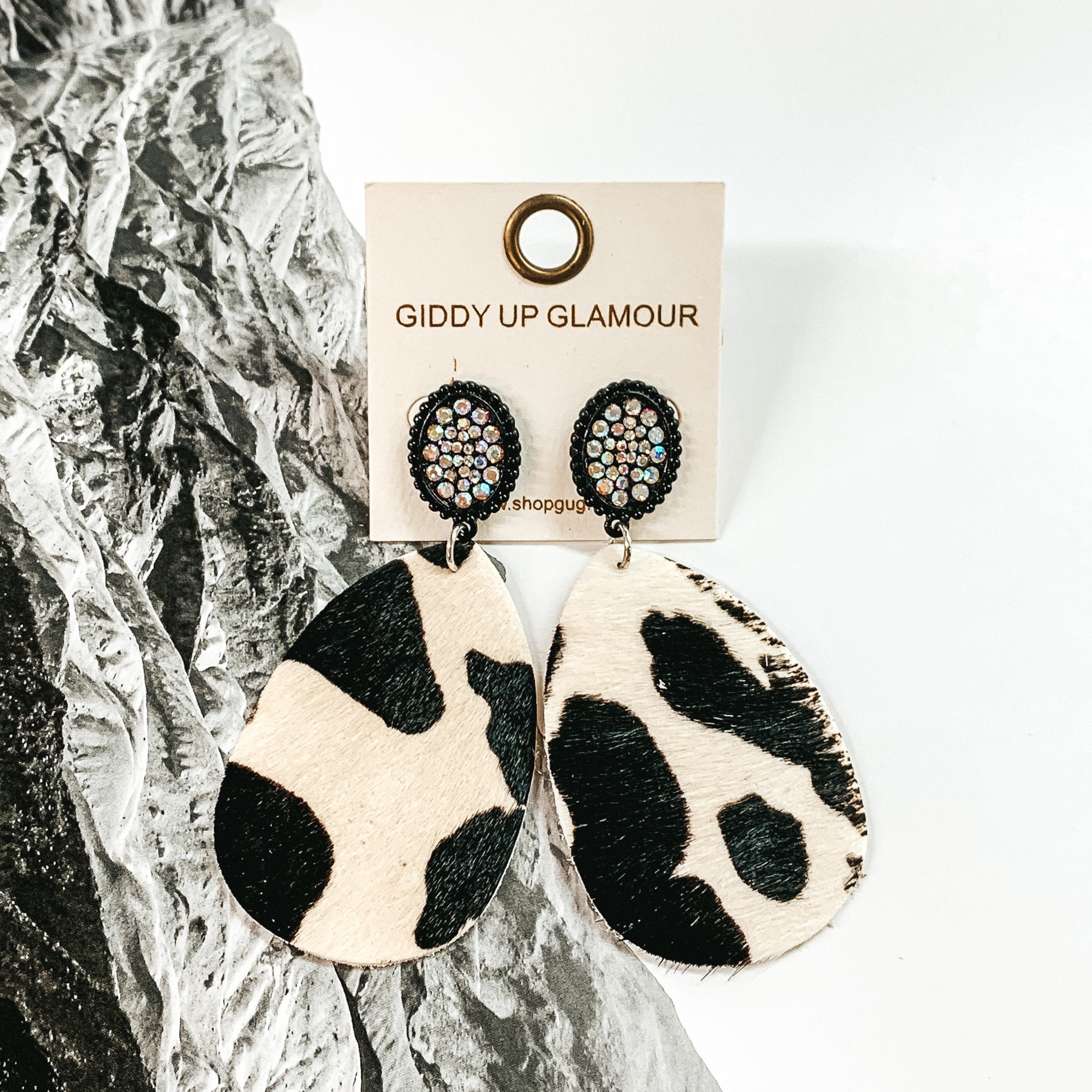 Black oval studs with AB crystals thrughout. These earrings also include a teardrop dangle that looks like a black and white cowhide. These earrings are pictured on a black and white picture of mountains. 