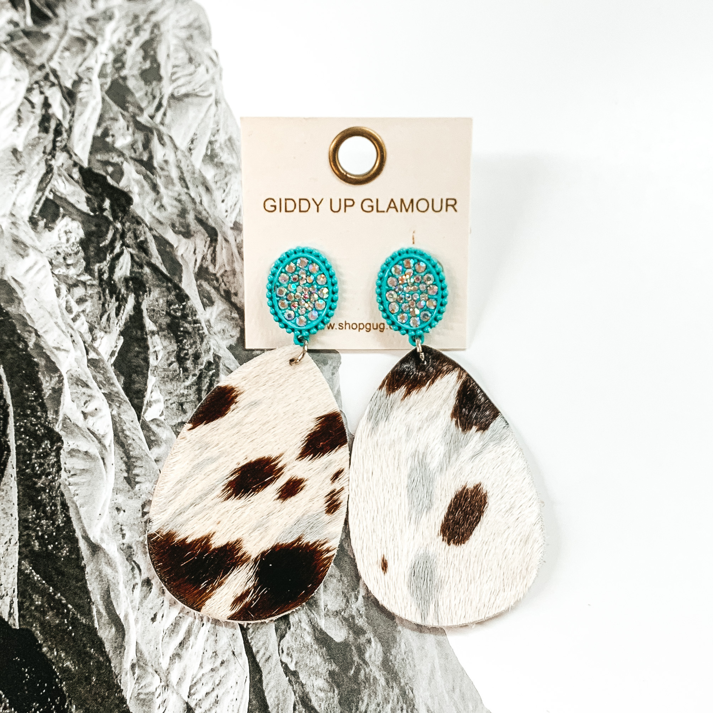 Turquoise oval studs with AB crystals thrughout. These earrings also include a teardrop dangle that looks like a brown, grey, and white cowhide.  These earrings are pictured on a black and white picture of mountains. 