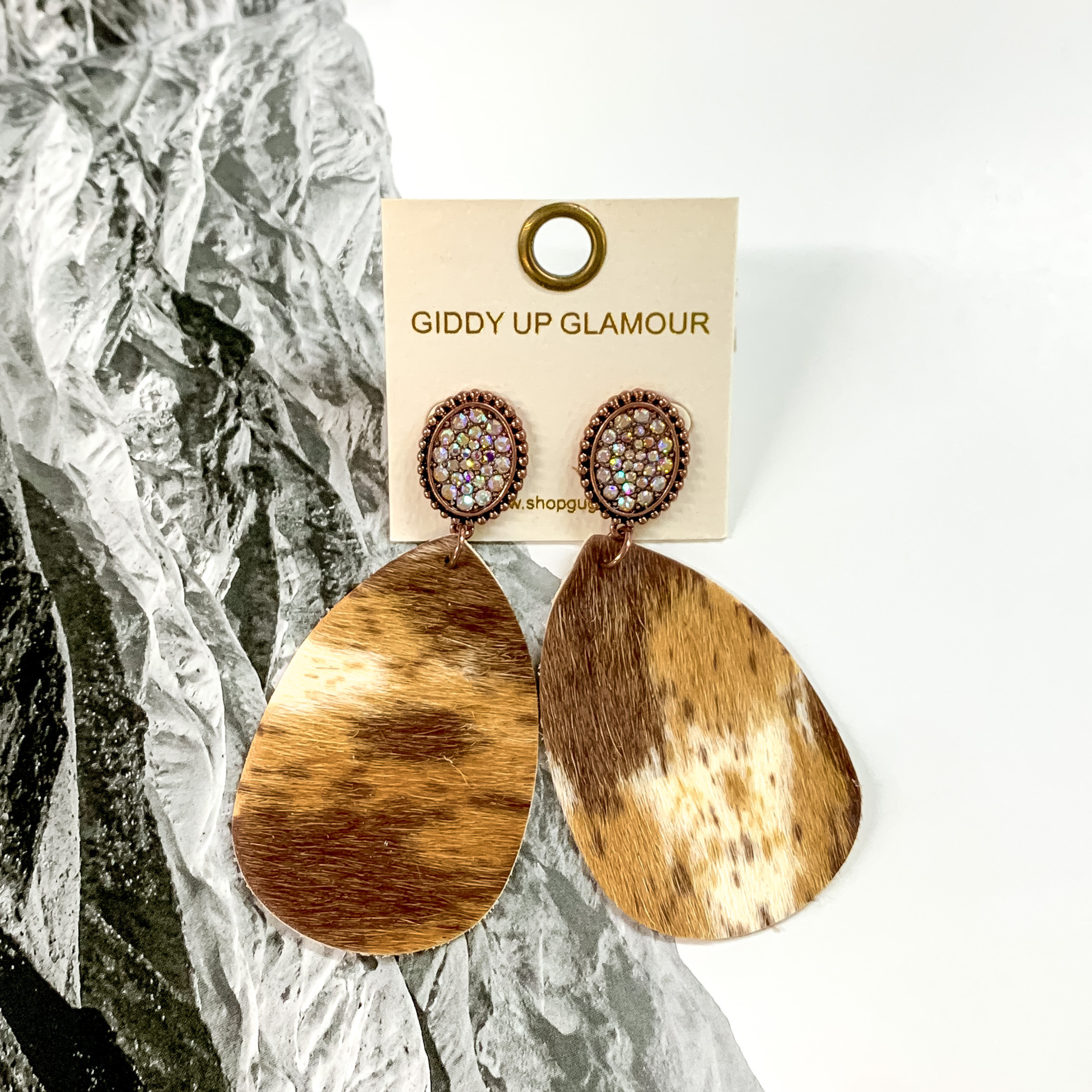 Copper oval studs with AB crystals thrughout. These earrings also include a teardrop dangle that looks like a brown, tan, and ivory cowhide.  These earrings are pictured on a black and white picture of mountains. 