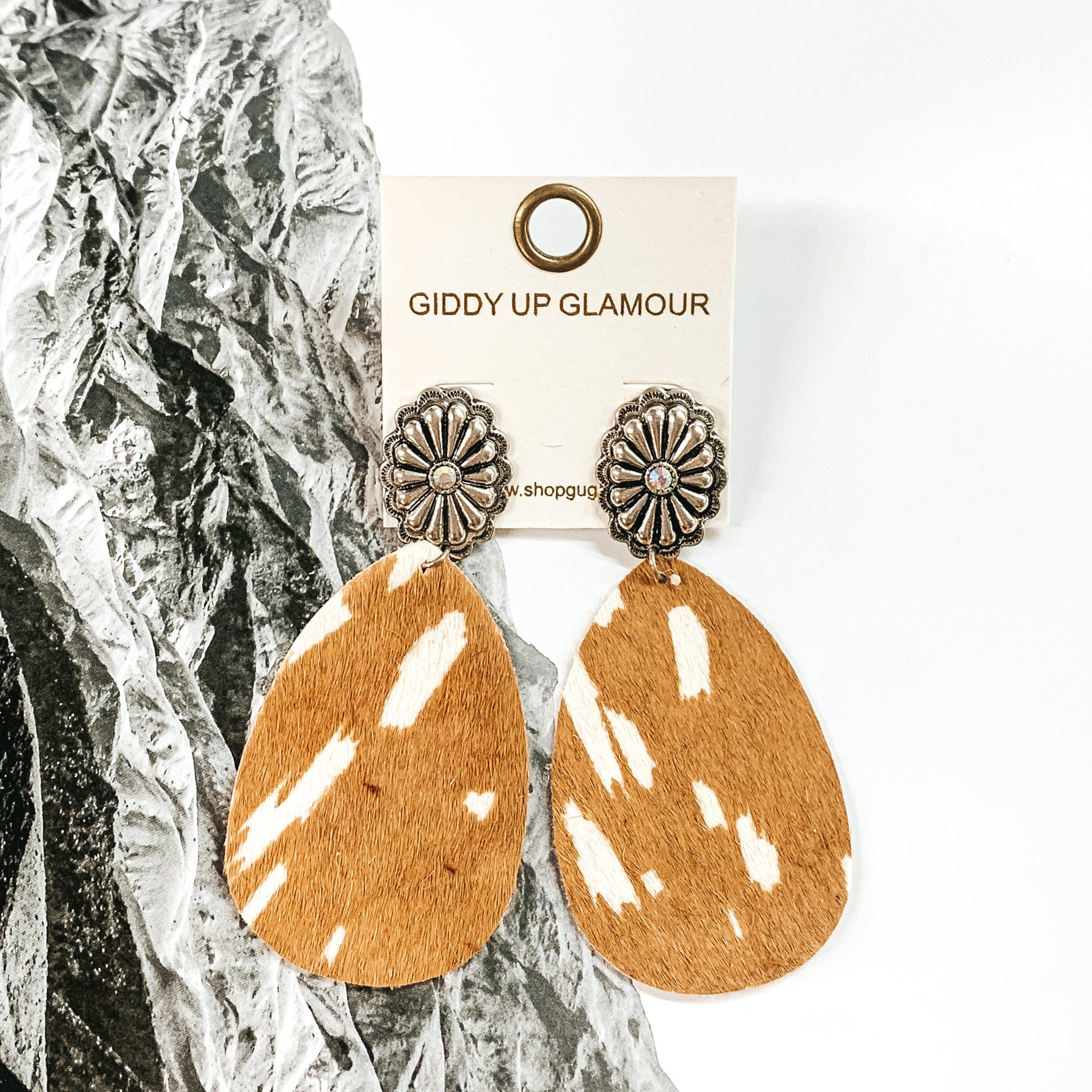 Silver oval concho studs with center AB crystal. These earrings also include a teardrop dangle that looks like a tan and ivory cowhide.  These earrings are pictured on a black and white picture of mountains. 