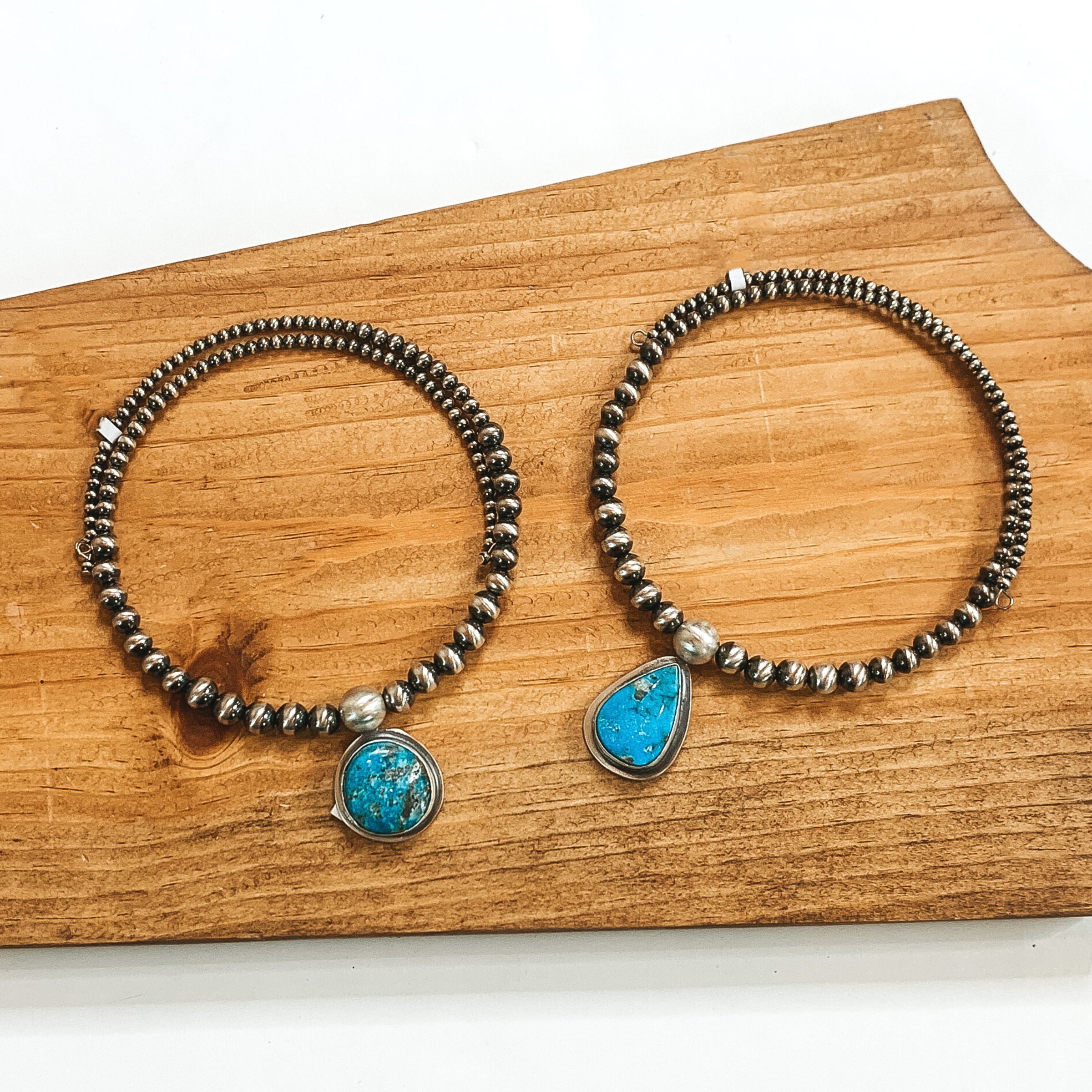 Two silver beaded wrap necklaces that each have a different shaped turquoise colored stone in the center of the necklace. These necklaces are pictured on a wood block on a white background. 