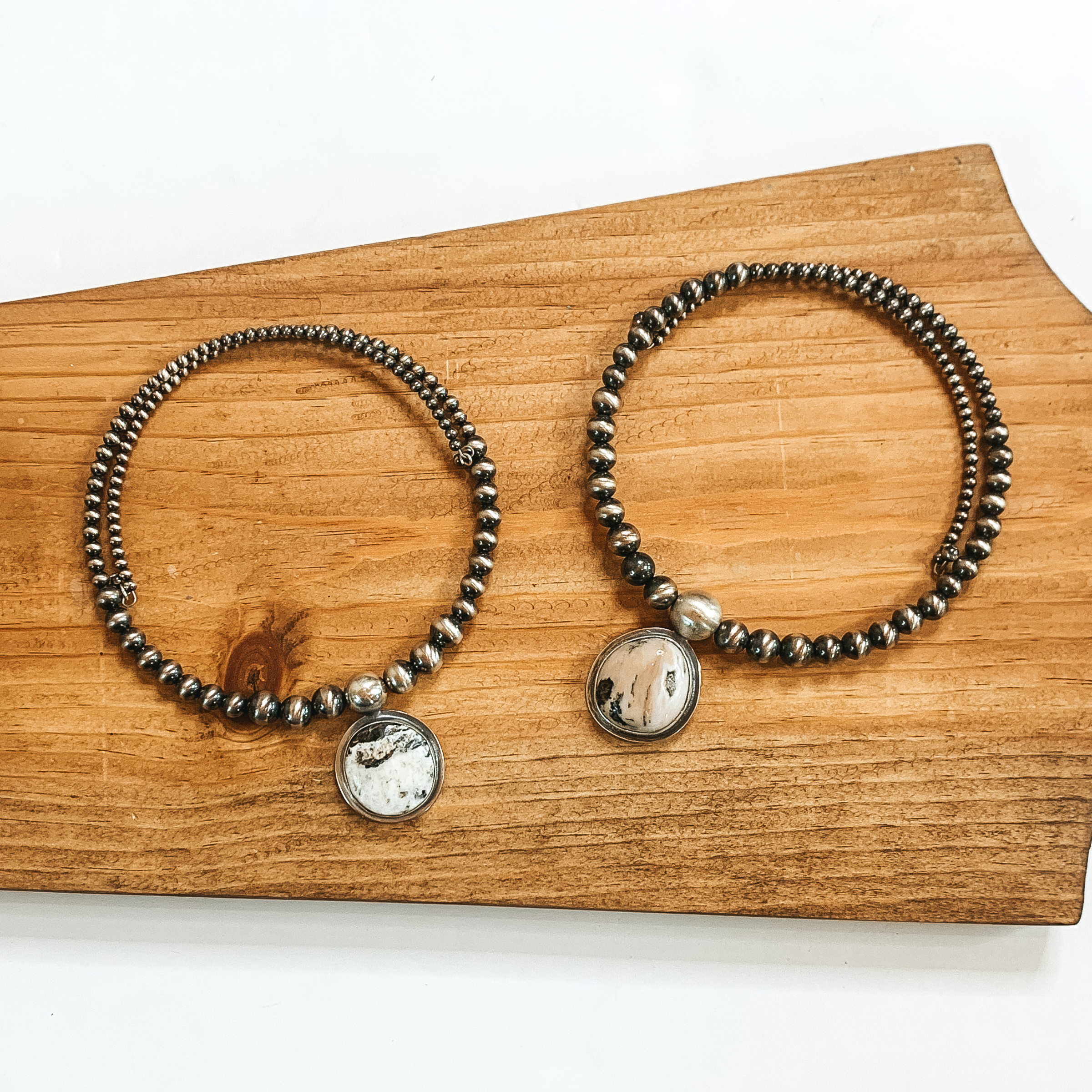 Two silver beaded wrap necklaces that each have a different shaped white buffalo colored stone in the center of the necklace. These necklaces are pictured on a wood block on a white background. 