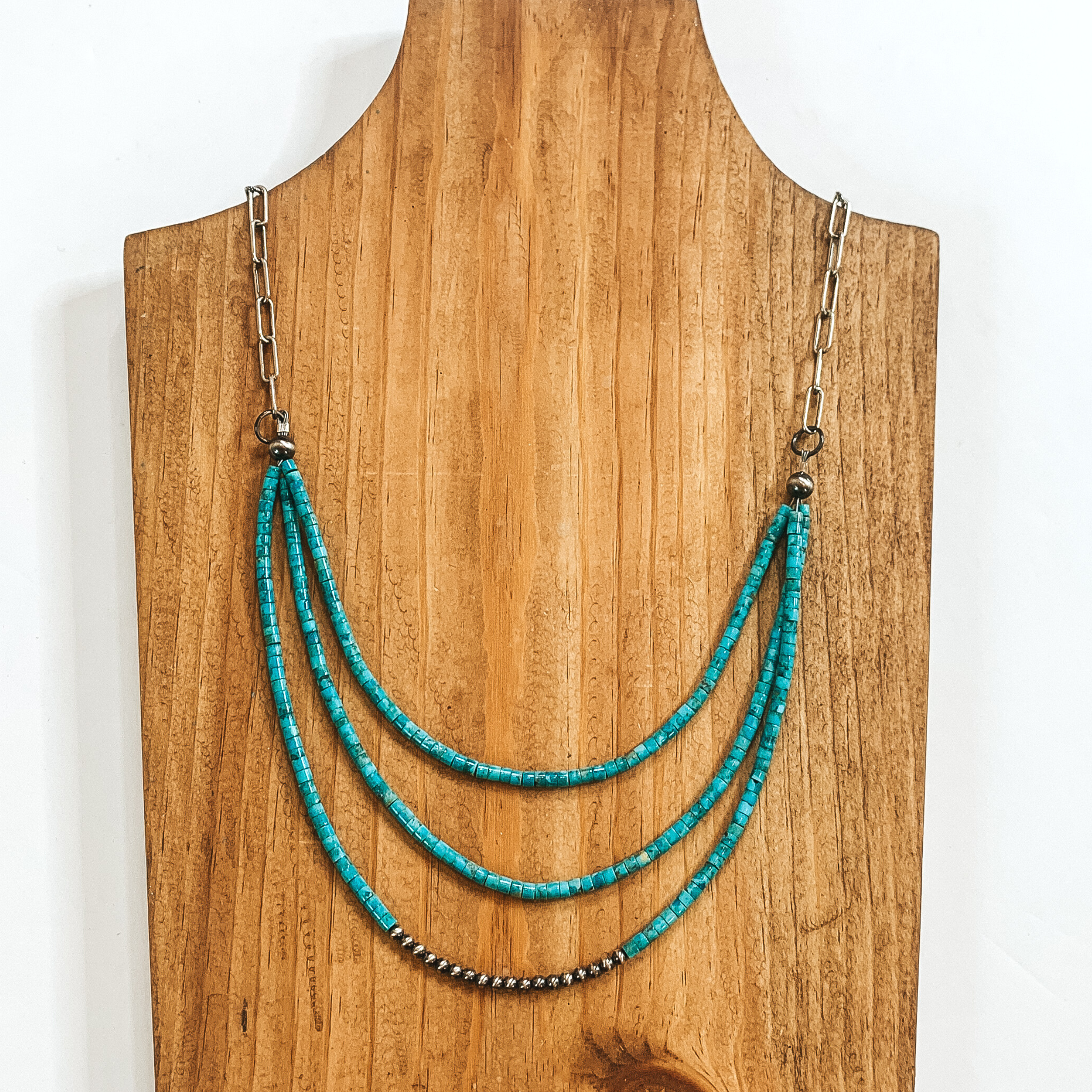 Silver paperclip chain with three turquoise beaded strands. The bottom strand also has a segment of silver beads. This necklace is pictured on a wood necklace holder on a white background. 