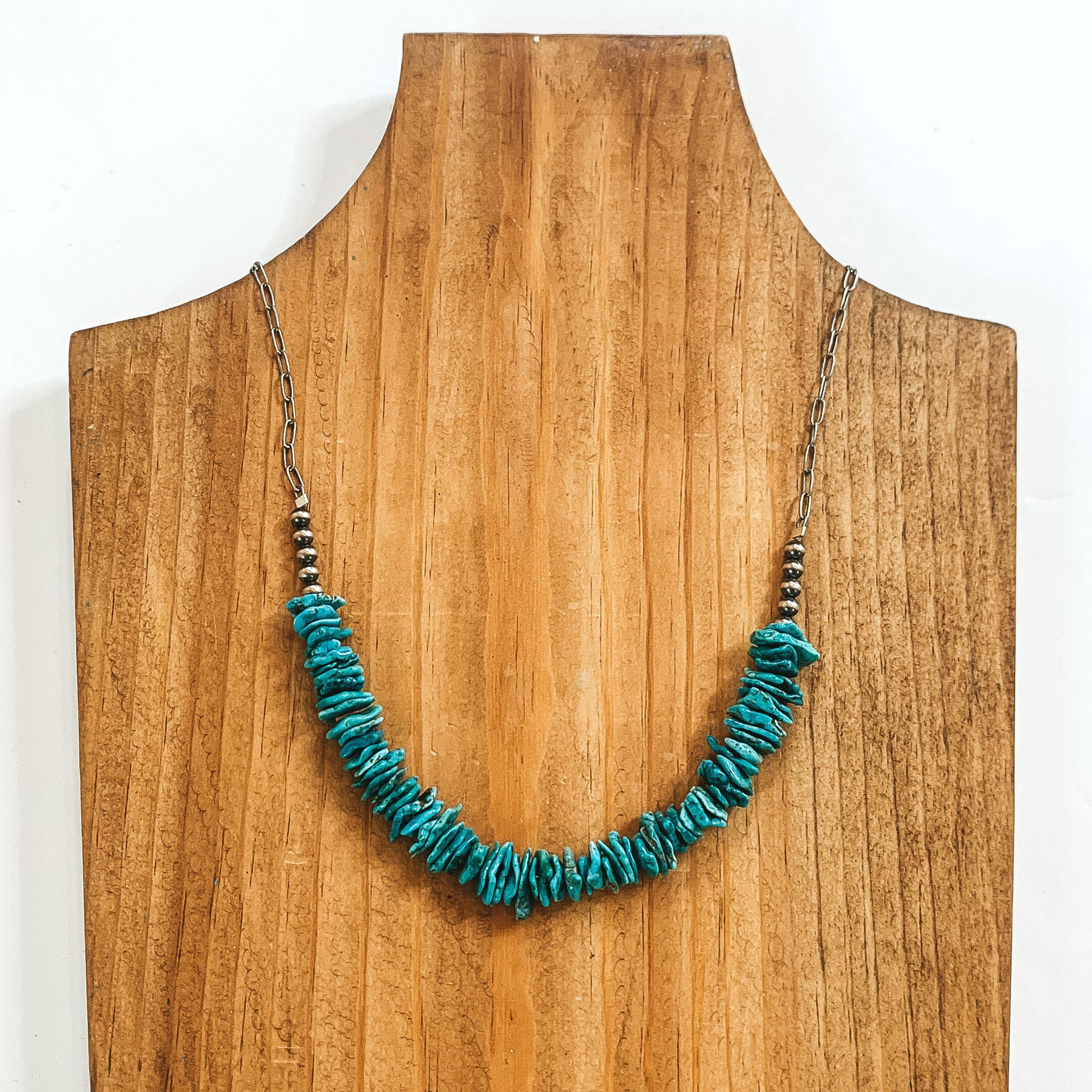 Thin paperclip chain with flat, turquoise beads on the bottom half. This necklace is pictured on a wood necklace holder on a white background. 