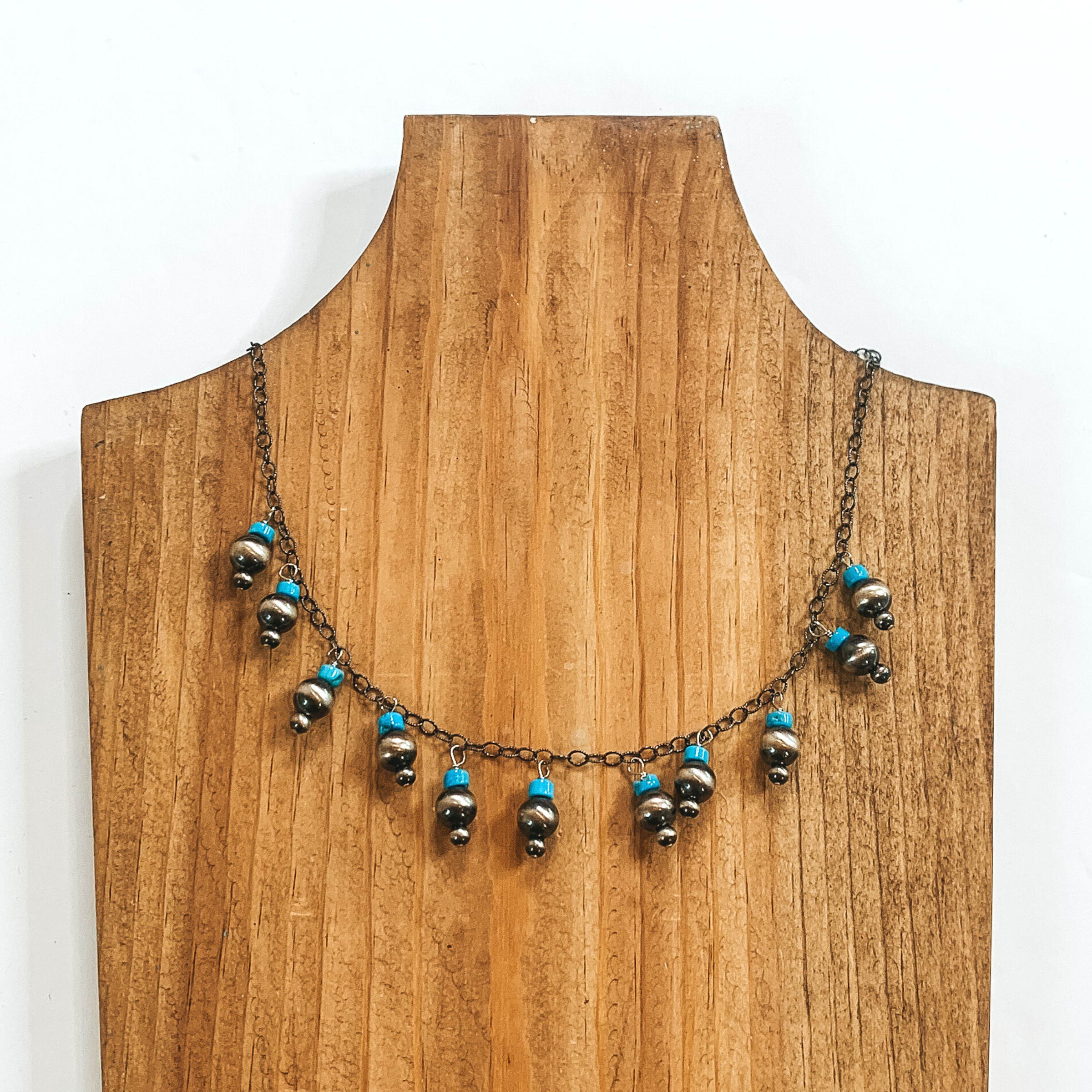 Silver chained necklace with charms that include a siver bead and turquoise bead stacked on each other. This necklace is pictured on a wood necklace holder on a white background. 