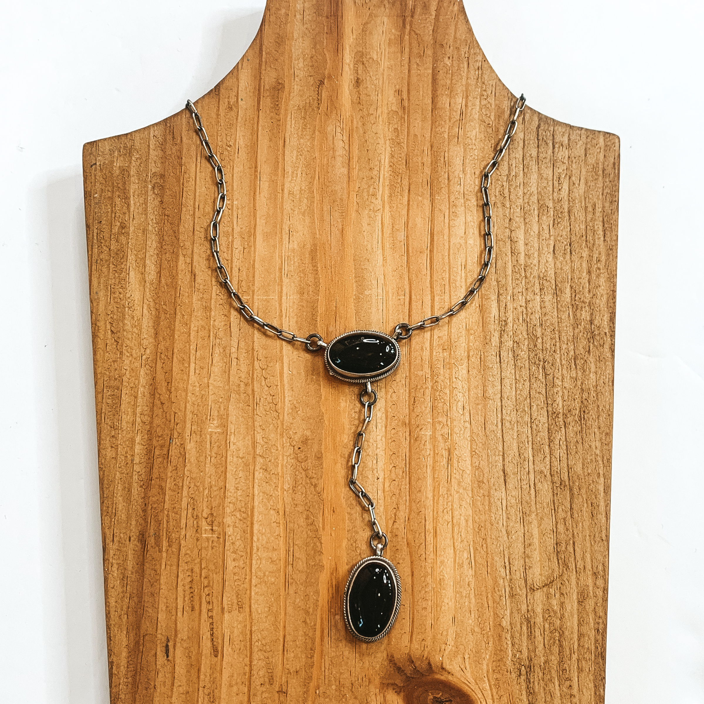 Silver chained necklace with oval black stone and a hanging black oval stone. This necklace is pictured on a wood necklace holder on a white background. 