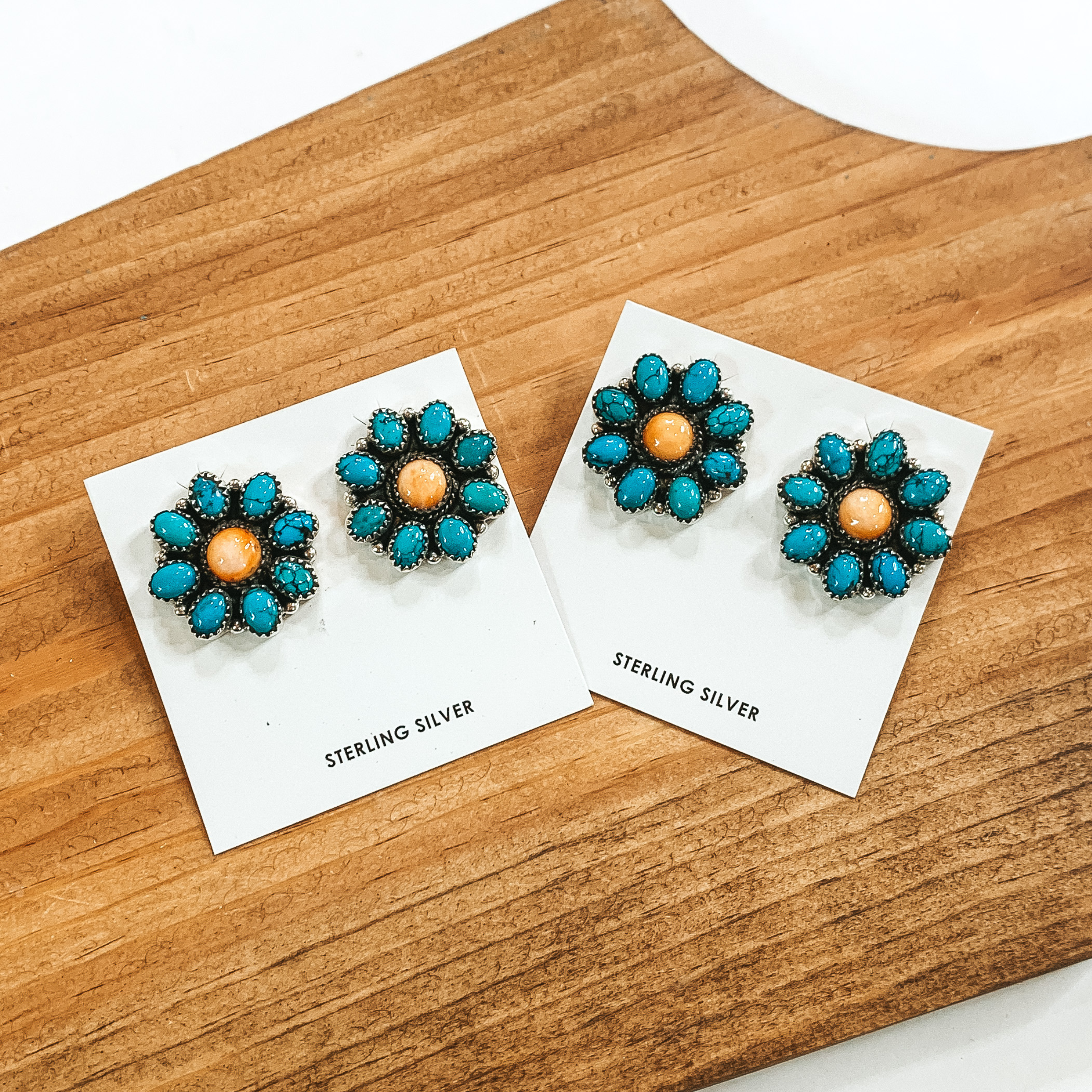 Turquoise stone flower cluster earrings that have an orange center stone. There are two pairs of these earrings pictured on a wood block on a white background. 