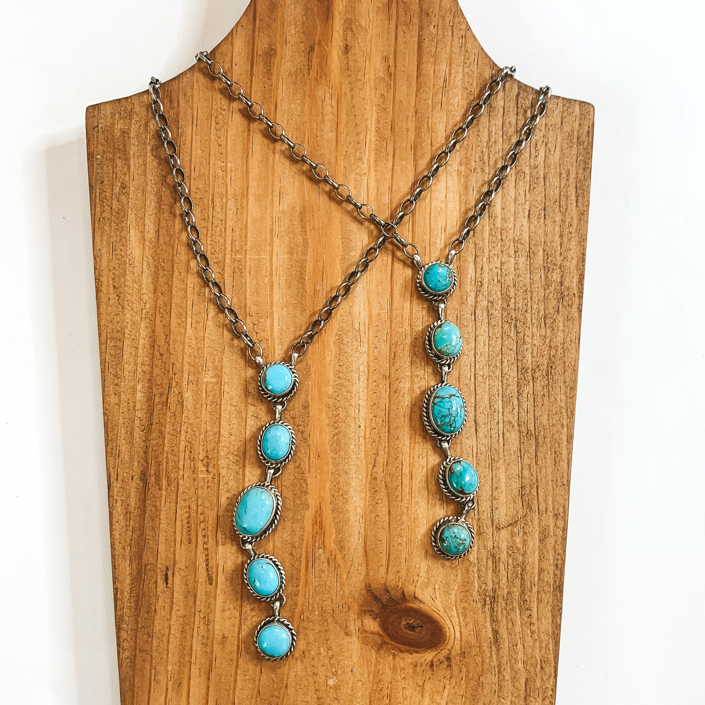 Paperclip chain necklace with five turquoise stone pendants hanging down from the center. There are two necklaces pictured on a wood necklace holder on a white background. 