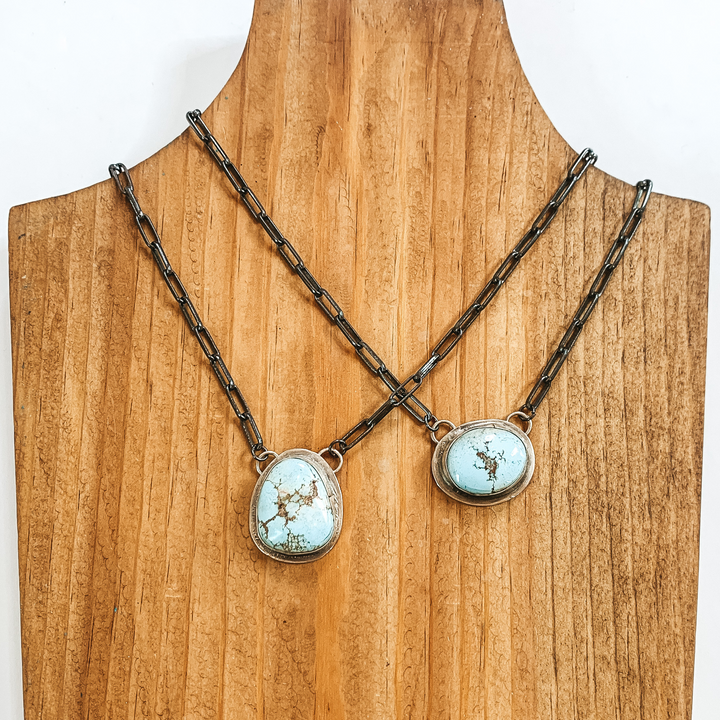 Paperclip chain necklace with a light blue center stone pendant. There are two necklaces pictured on a wood necklace holder on a white background. 