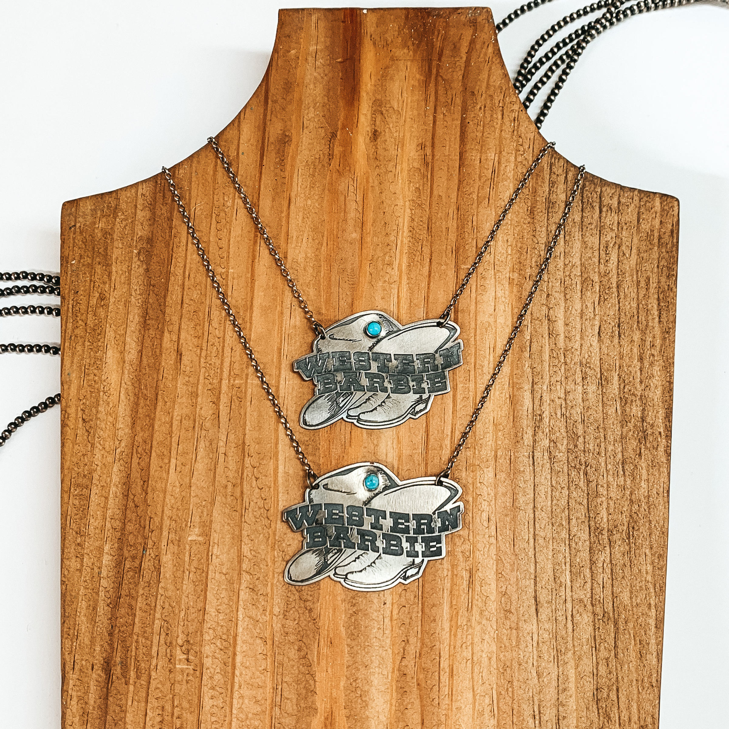 Silver chain necklace with a silver pendant in the shape of a hat and boots and small turquoise stone. The silver pendant has the words "WESTERN BARBIE". This necklace is pictured on a wood block on a white background. 