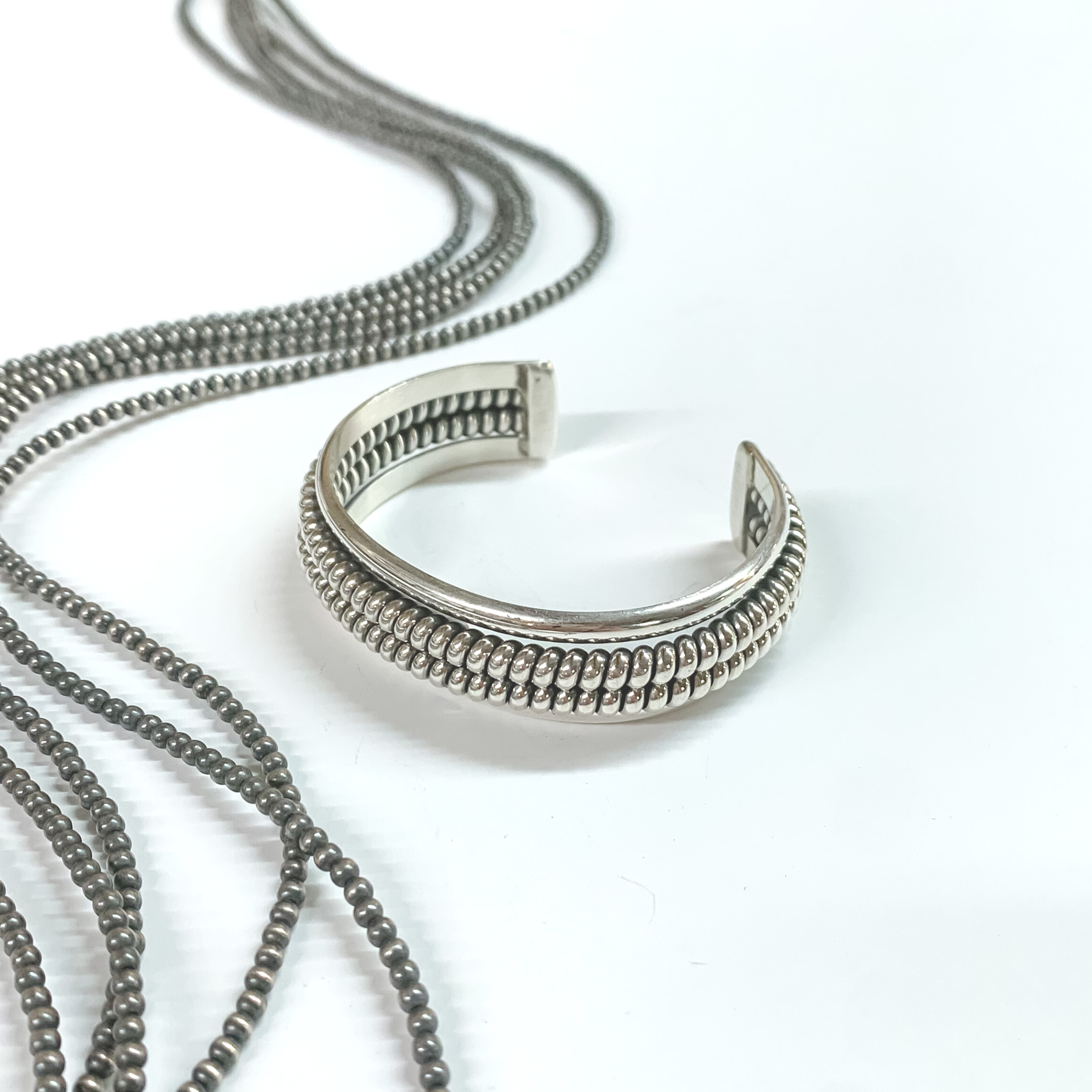 Silver cuff bracelet with a rope design. The rope design has two layers. This bracelet is pictured on a white background with silver beads on the left side. 