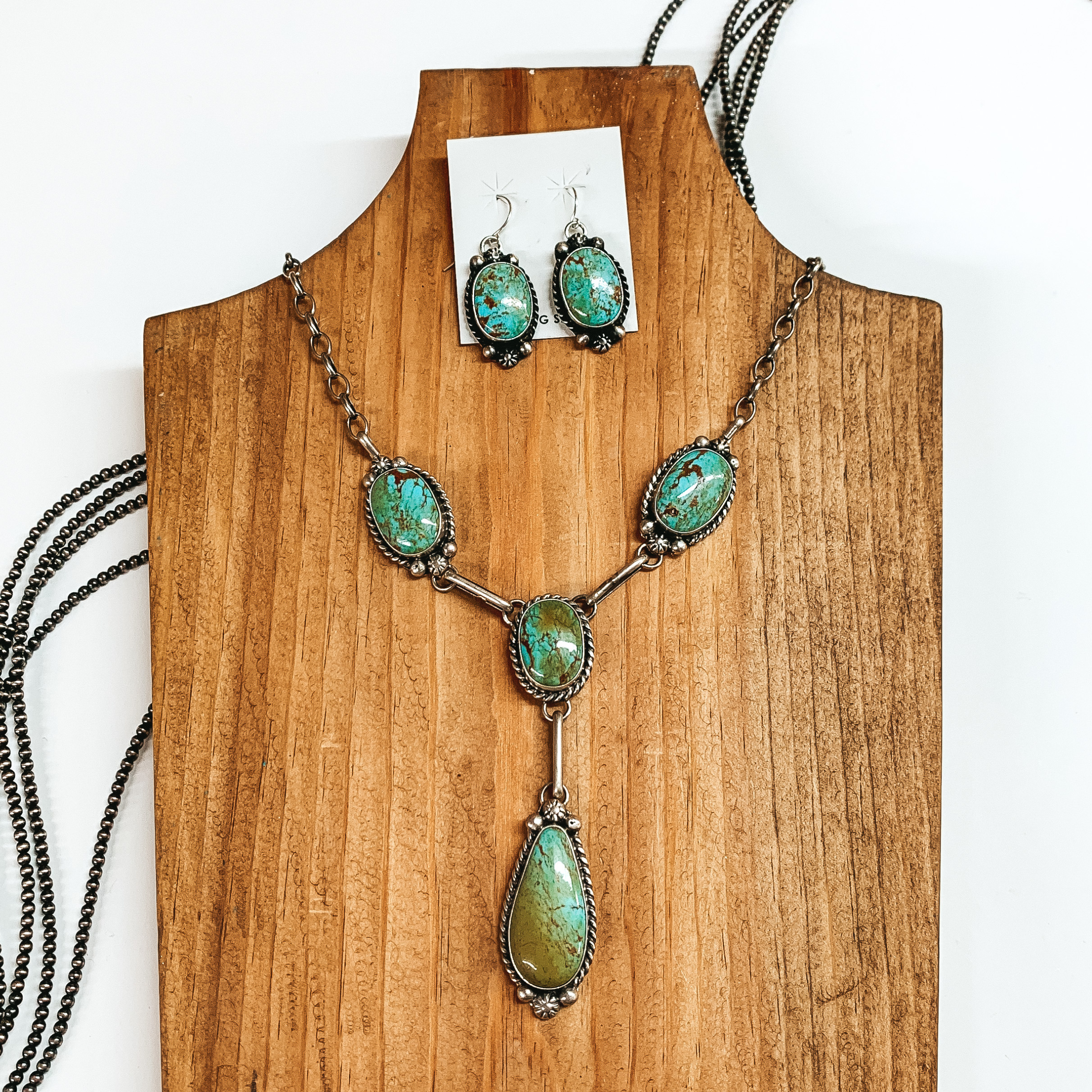 Silver lariat necklace with four turquoise stones pictured on a wood necklace holder. This picture also includes a pair of earrings that look like the stones on the necklace. This is all pictured on a white background. 