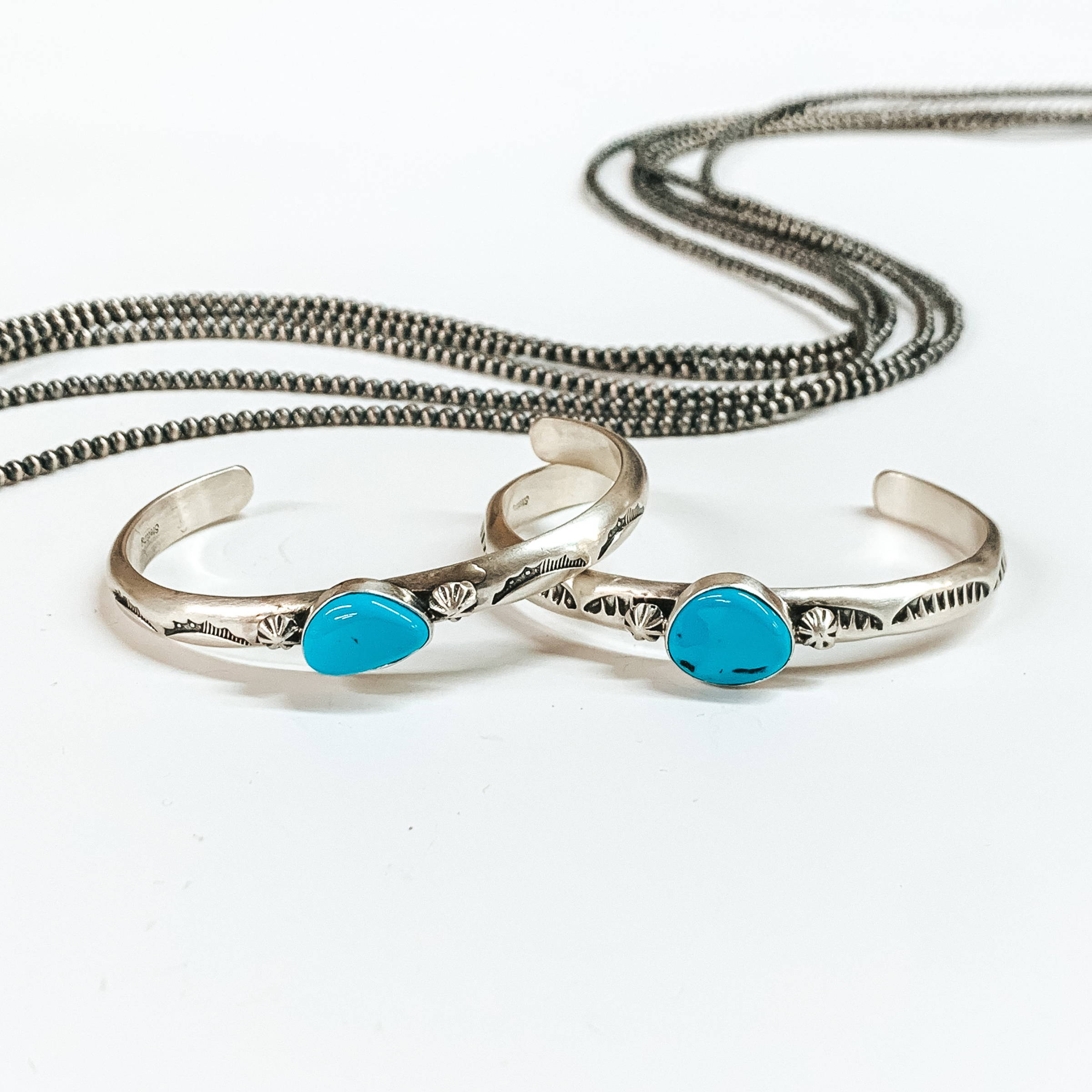 Two silver bangle bracelet with center turquoise stones. These bangles also include engraved details. These bracelets are pictured on a white background with silver beads at the top of the picture. 